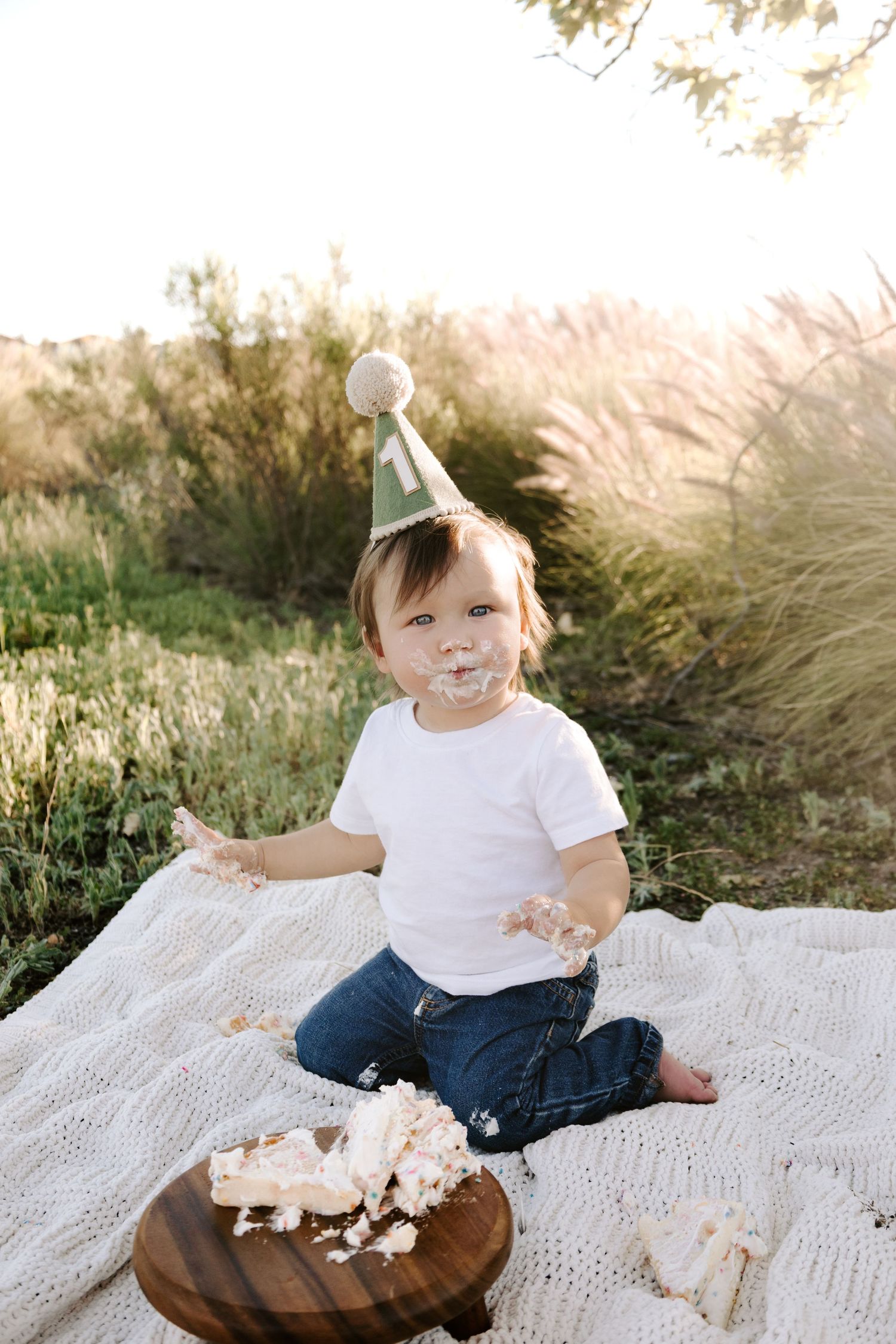 Happy baby in green party hat celebrates first birthday with cake smash in sunny field.