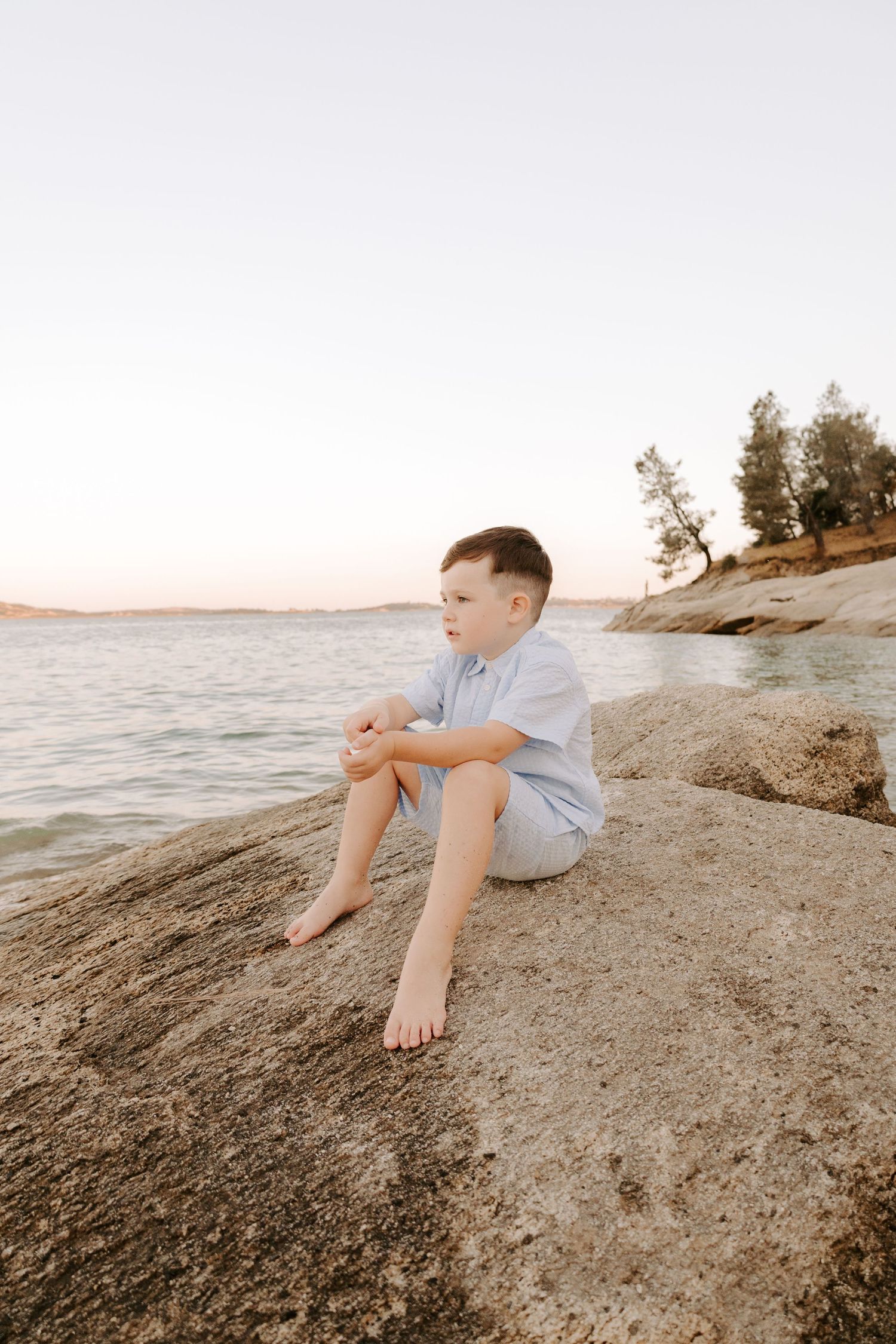 Young barefoot boy sits alone on rocky lakeshore gazing at the water during sunset.