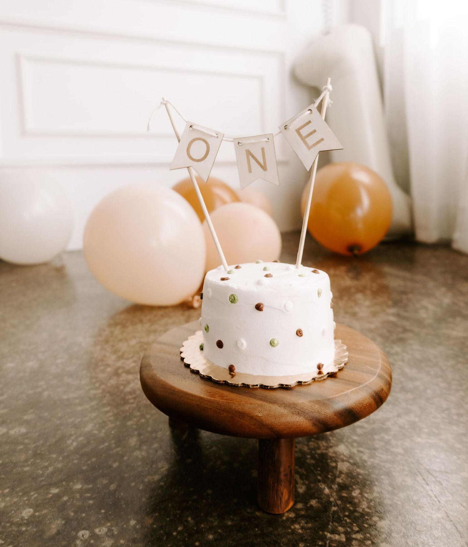 White first birthday cake with ONE banner topper on wood stand surrounded by balloons.