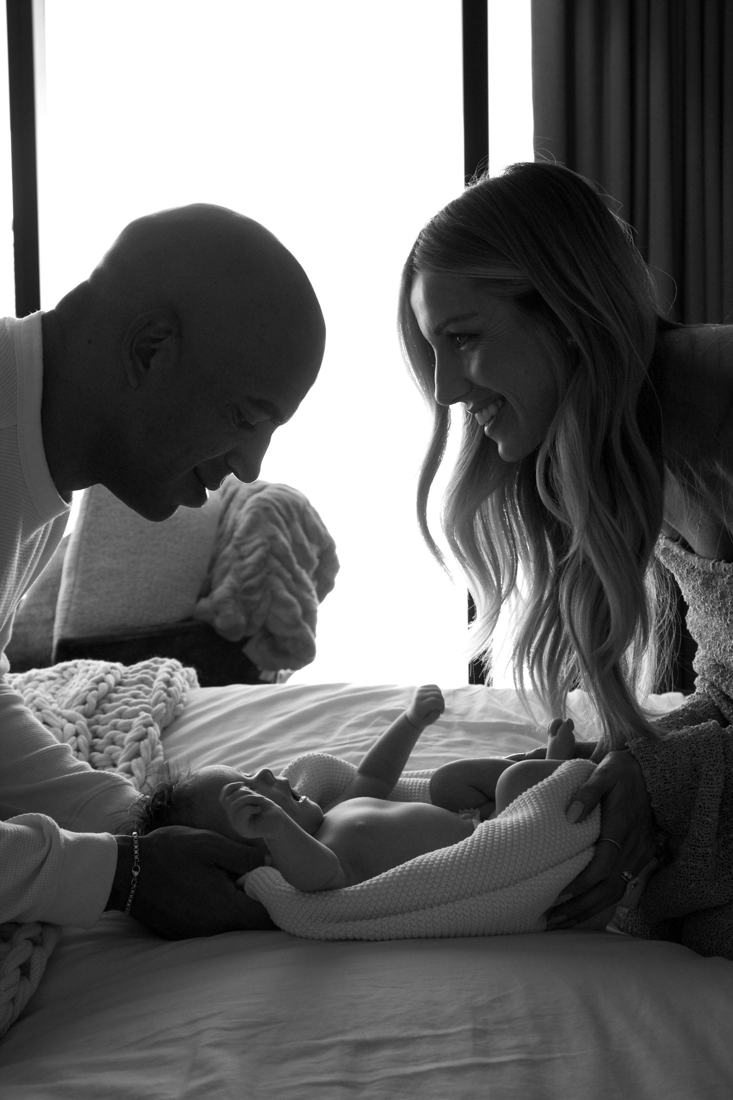 Black and white photo of two parents smiling lovingly at their newborn baby lying on a bed.