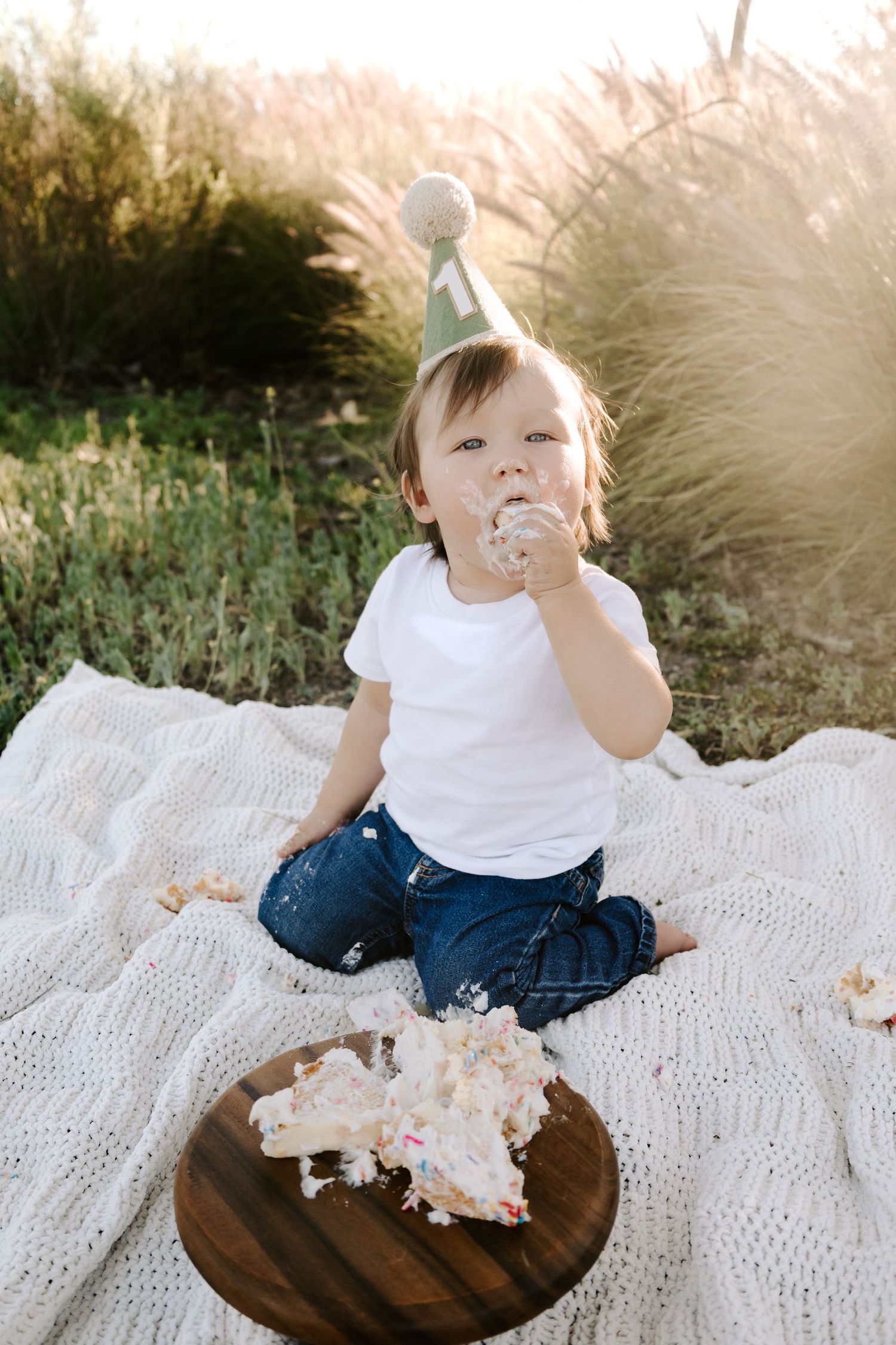 A one-year-old in a birthday hat sits on a blanket eating cake at a golden hour outdoor celebration.