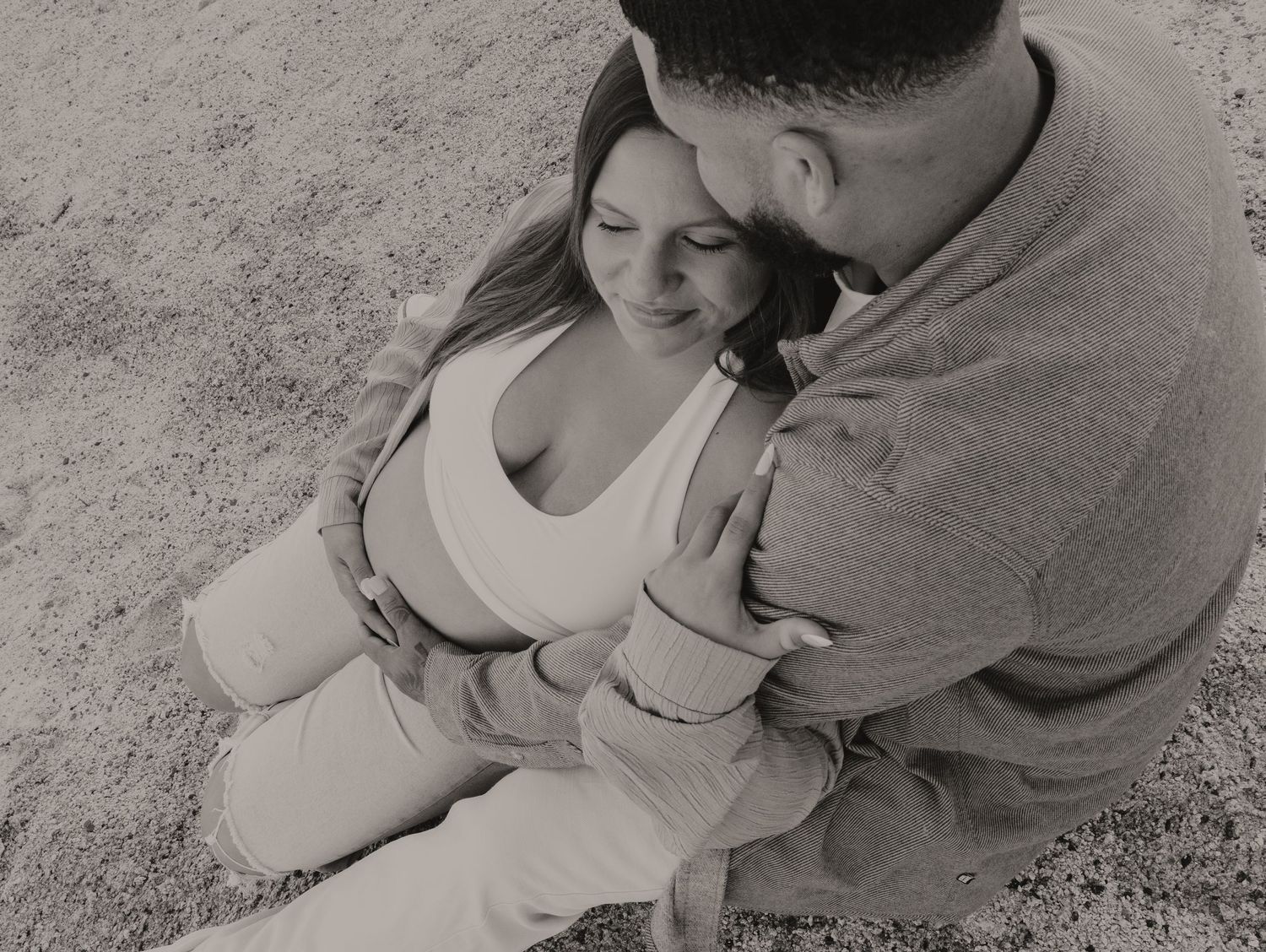 Black and white overhead photo of a couple sitting closely together on sandy ground outdoors.
