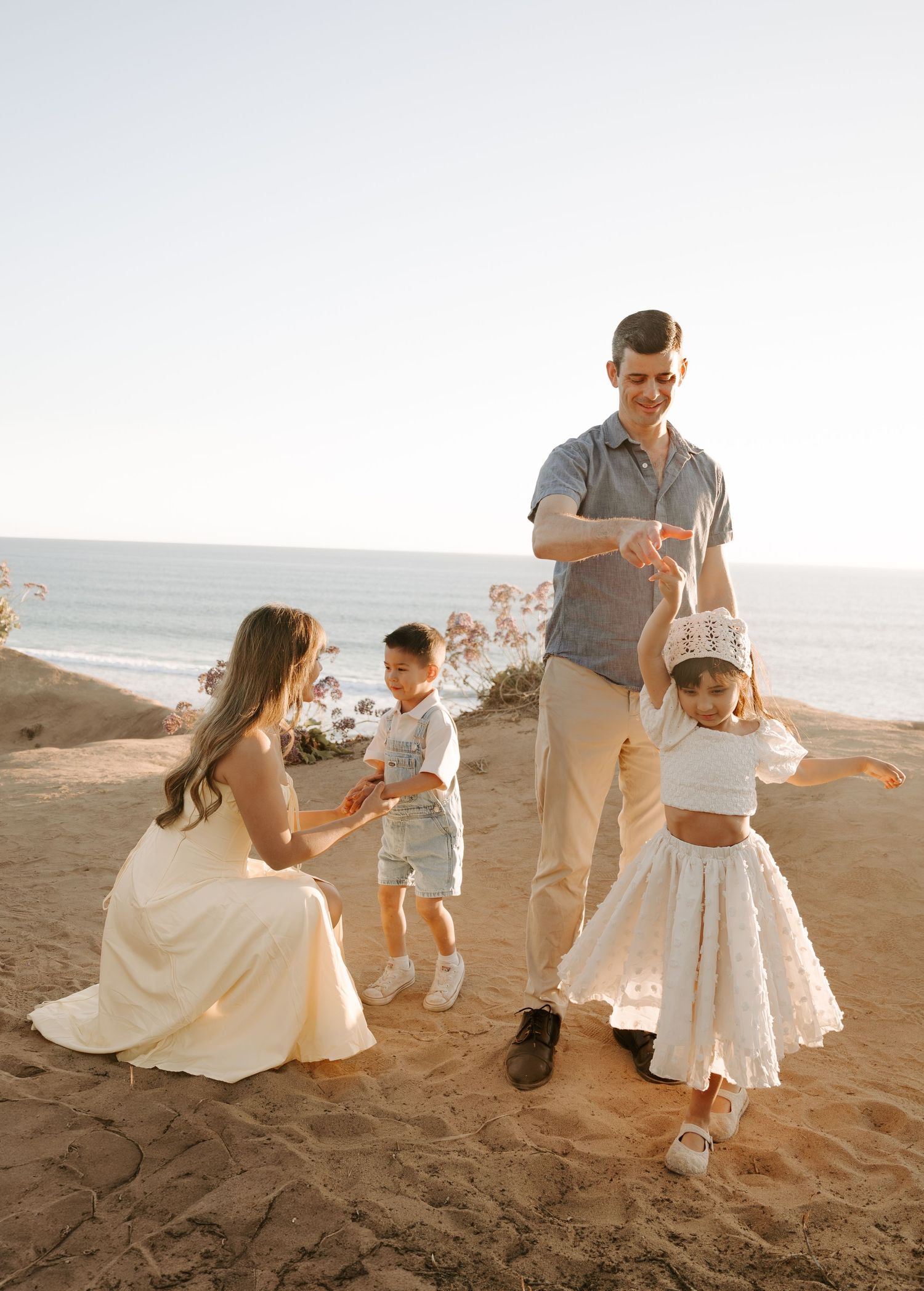 Family of four enjoying a sunny beach day on sandy cliffs, with children dancing near the ocean.
