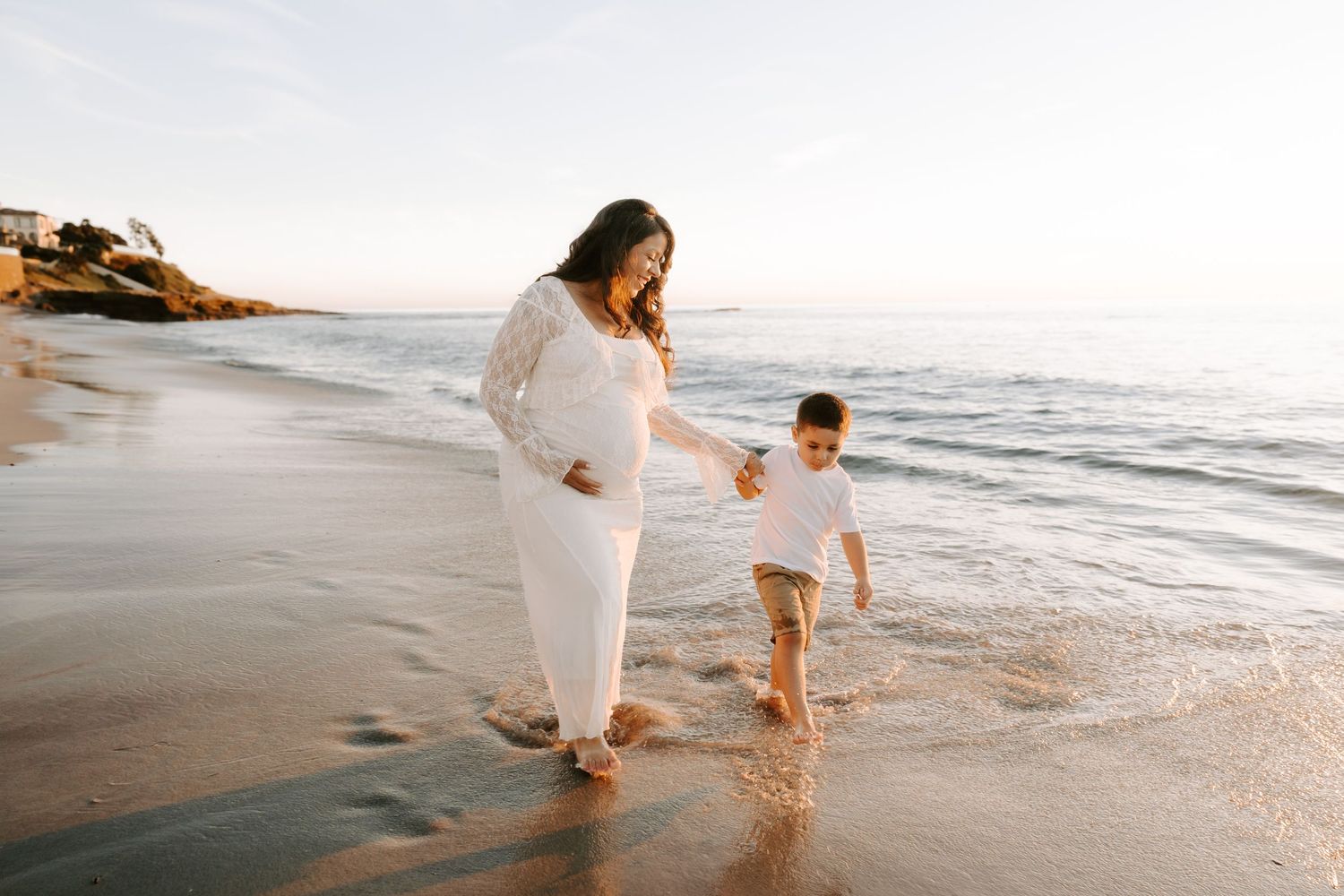 Pregnant woman in white lace dress walks hand-in-hand with young boy along sunny beach shoreline.