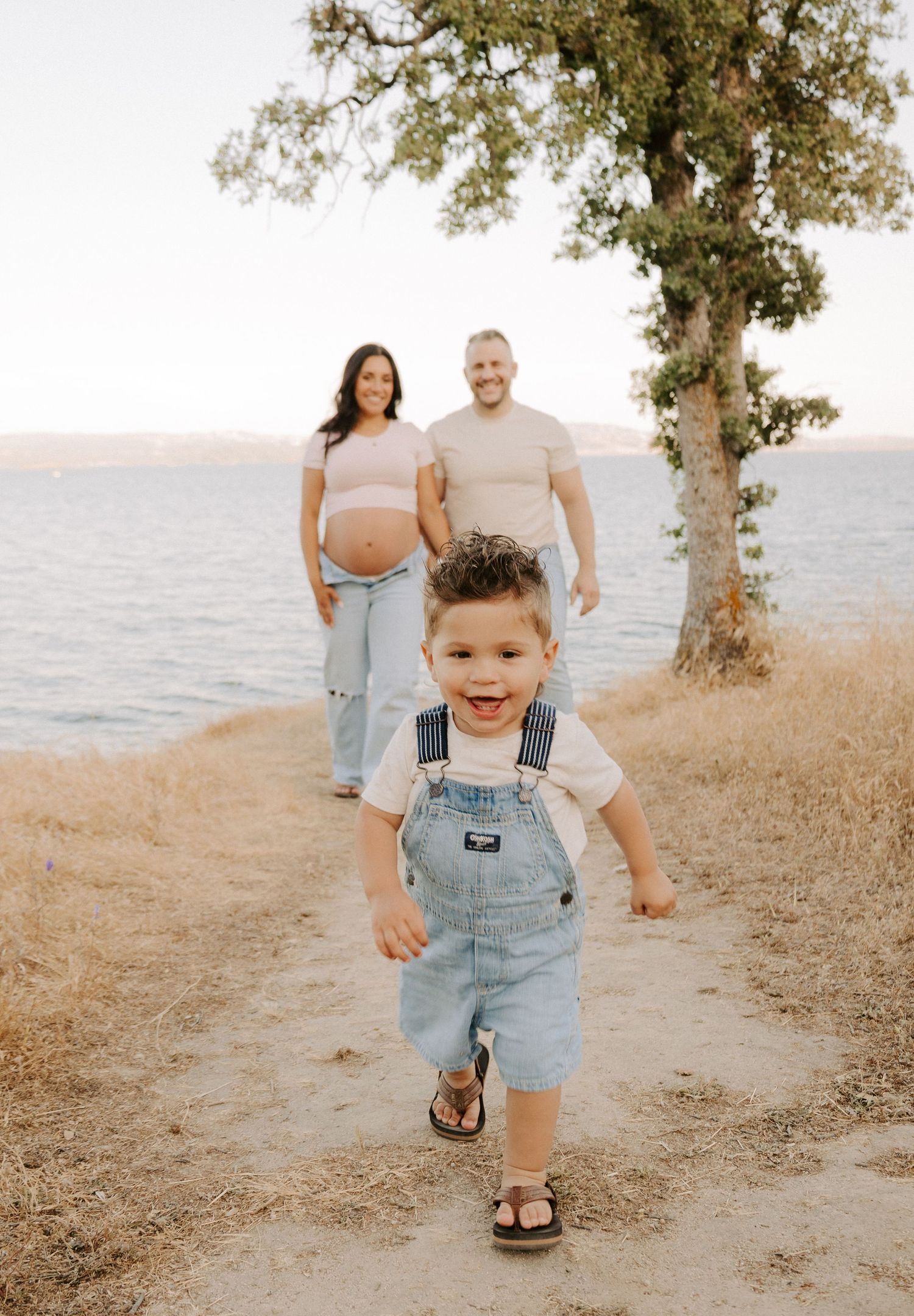 Excited toddler in denim overalls runs toward camera while expecting parents smile in background near lake.