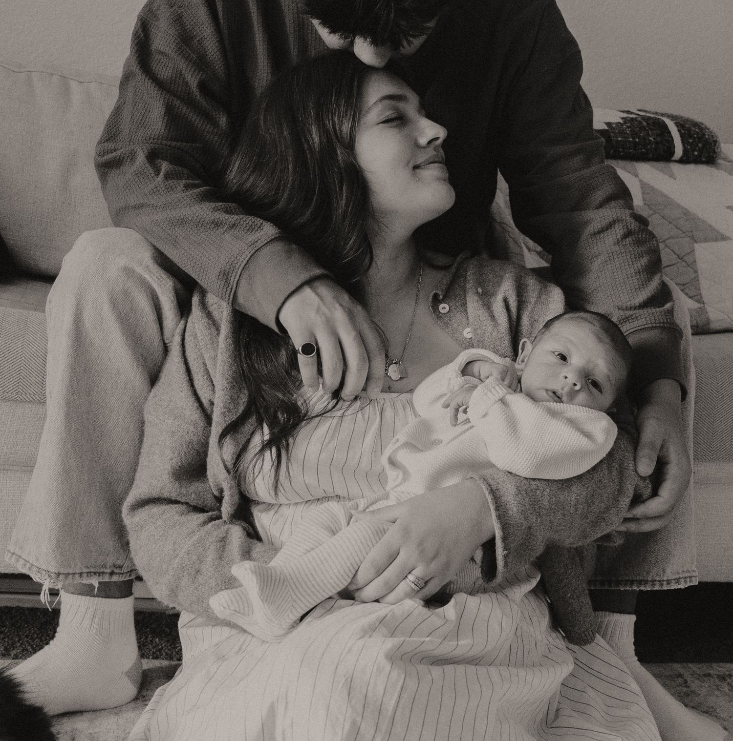 Black and white intimate portrait of new parents cradling their newborn baby on a cozy couch.