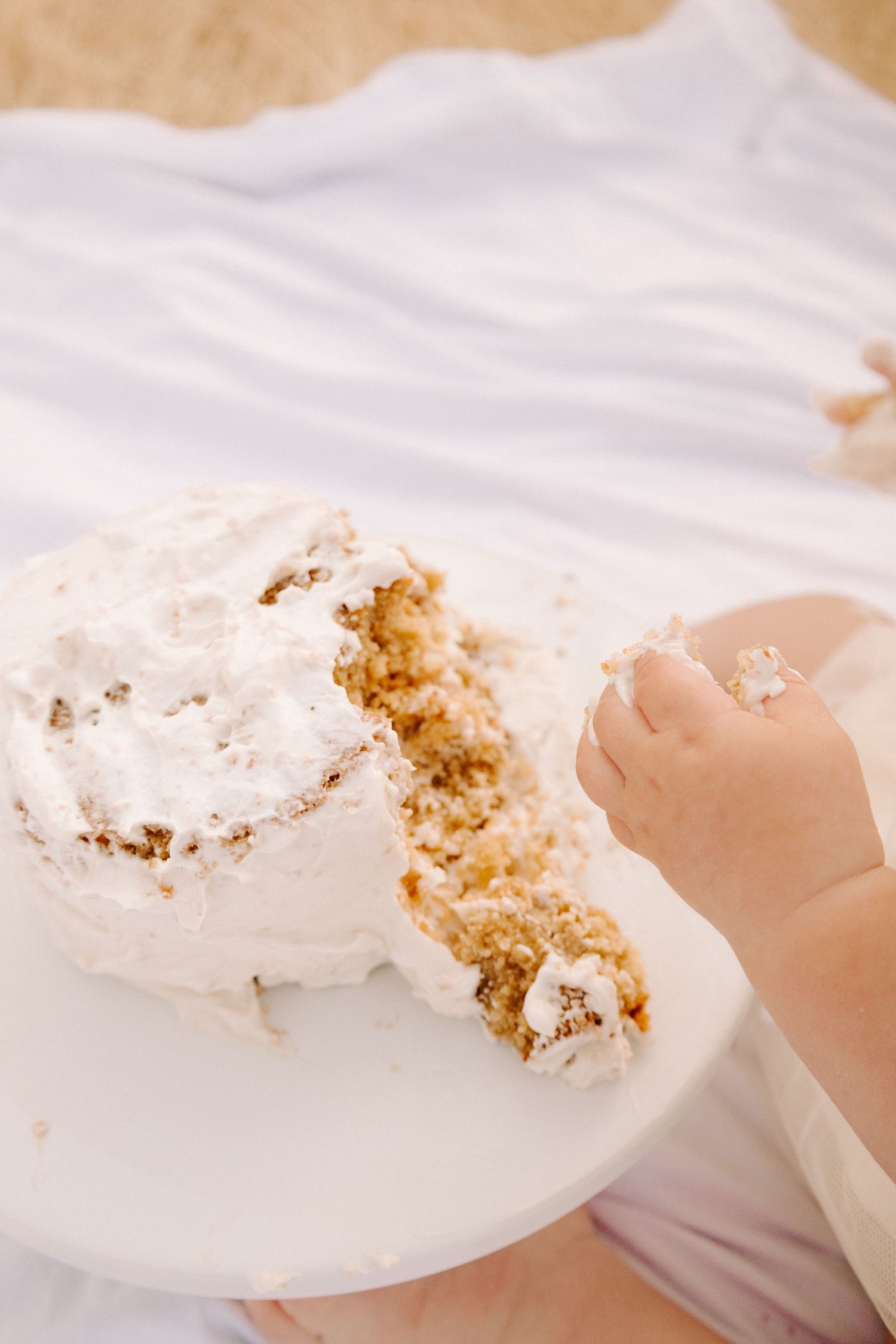 Close-up of baby hands pulling apart white frosted smash cake on white cake stand during first birthday.