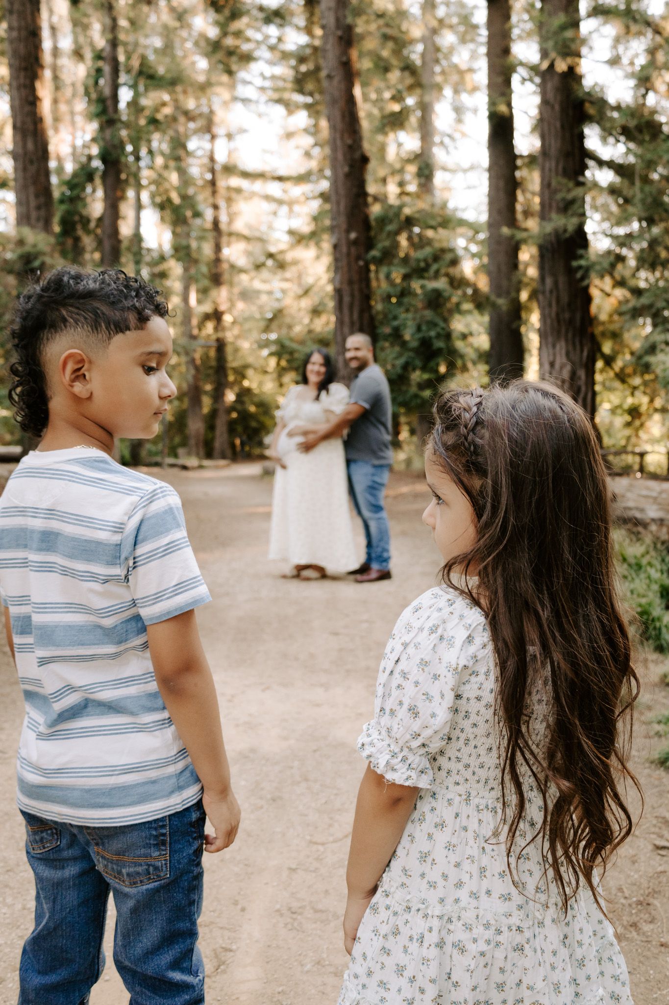 Two children stand on a forest path while their parents share a tender moment in the background during a maternity photoshoot.