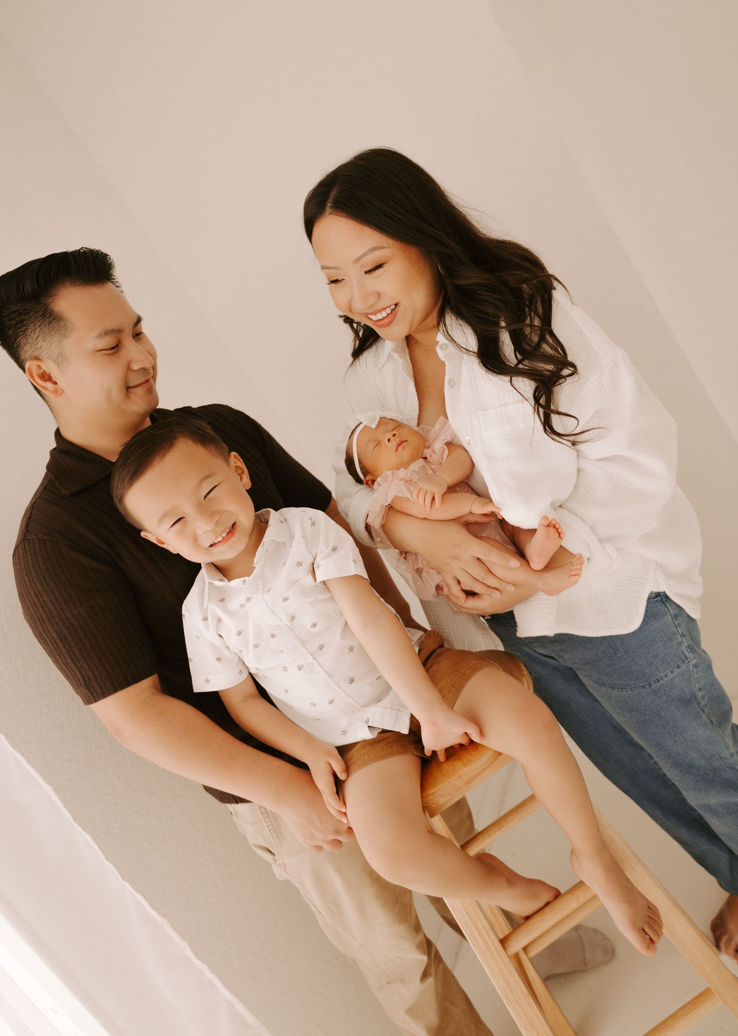 Happy Asian family of four poses in a bright studio, parents smiling as mom holds their newborn baby girl.