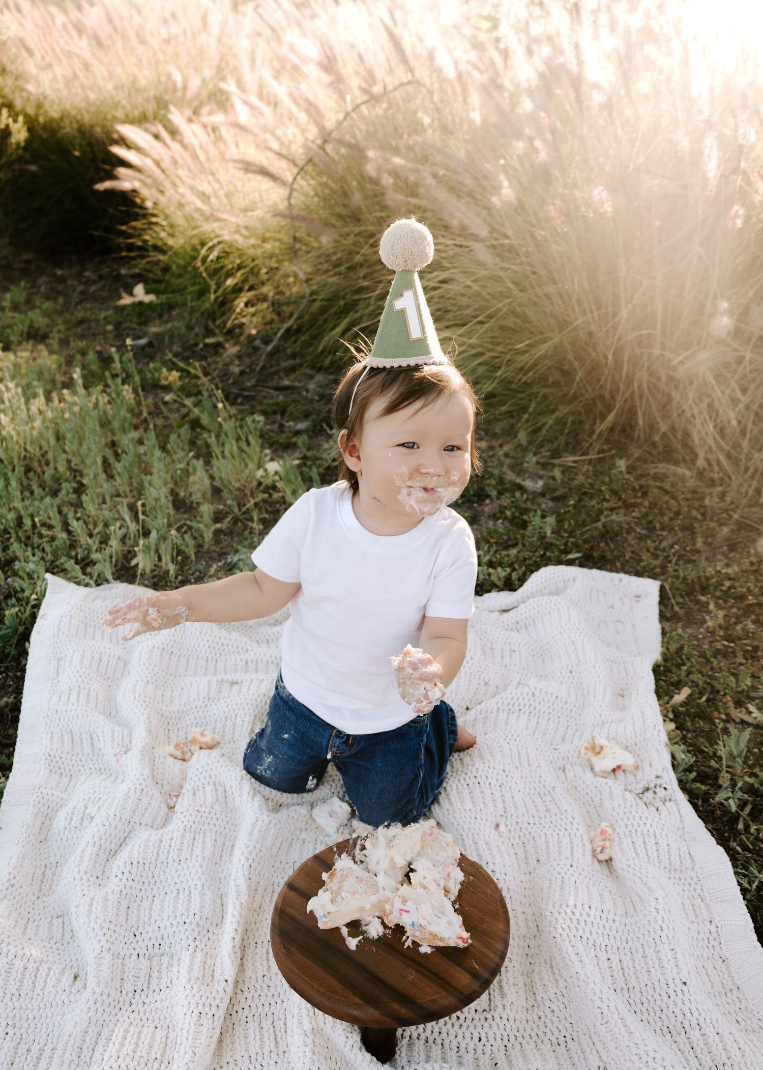 Toddler in birthday hat smashes first cake outdoors on white blanket in golden sunlight.