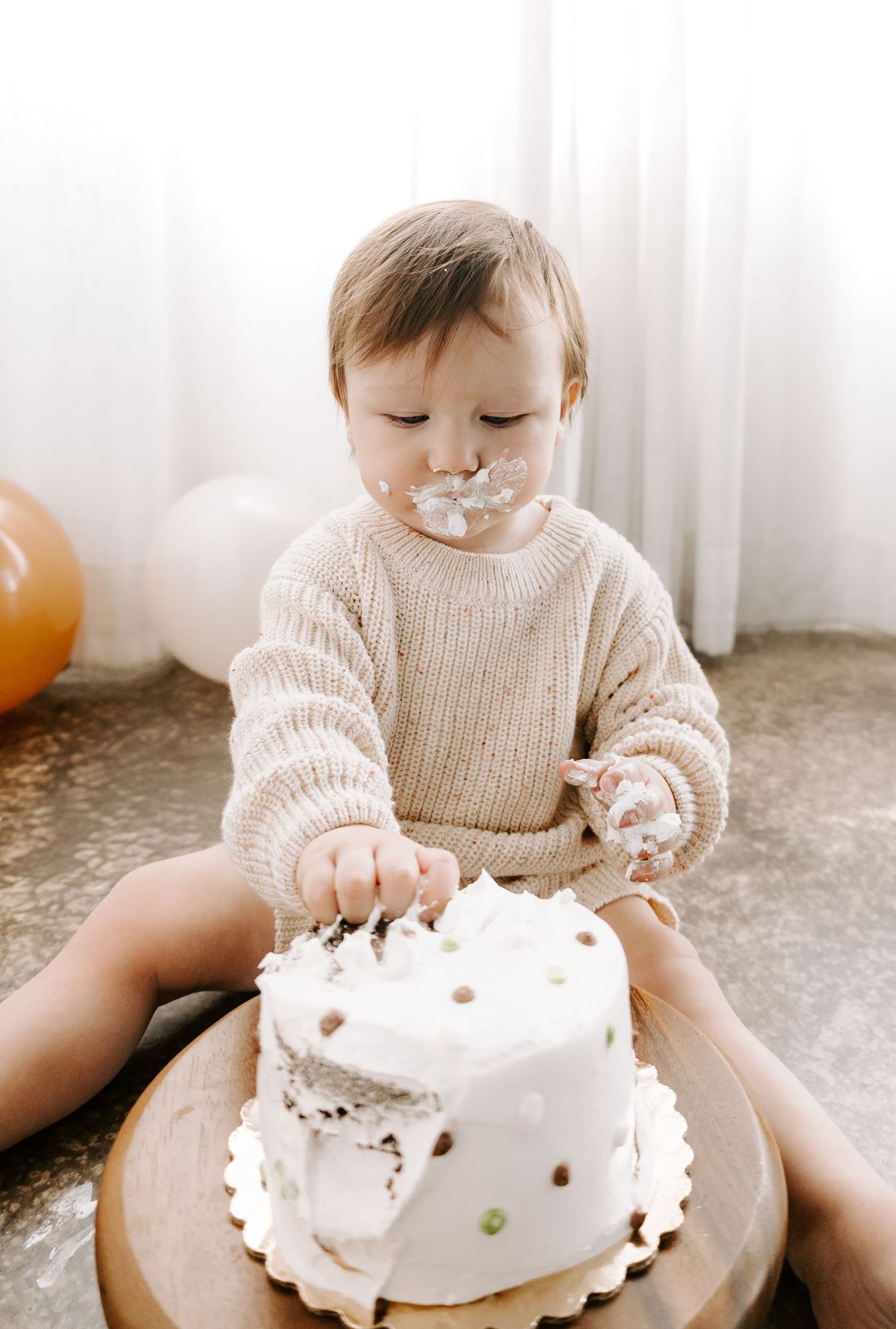 Baby in knit sweater eats first birthday cake decorated with polka dots and white frosting.