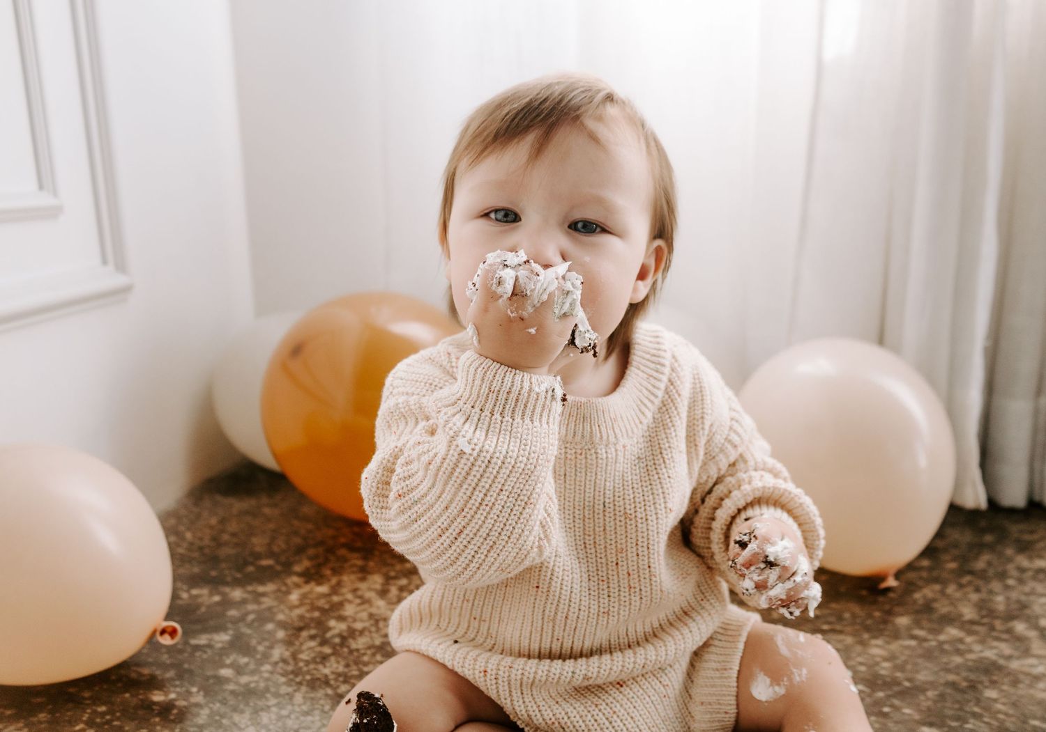 Adorable baby in beige sweater enjoys first birthday cake with orange and cream balloons.