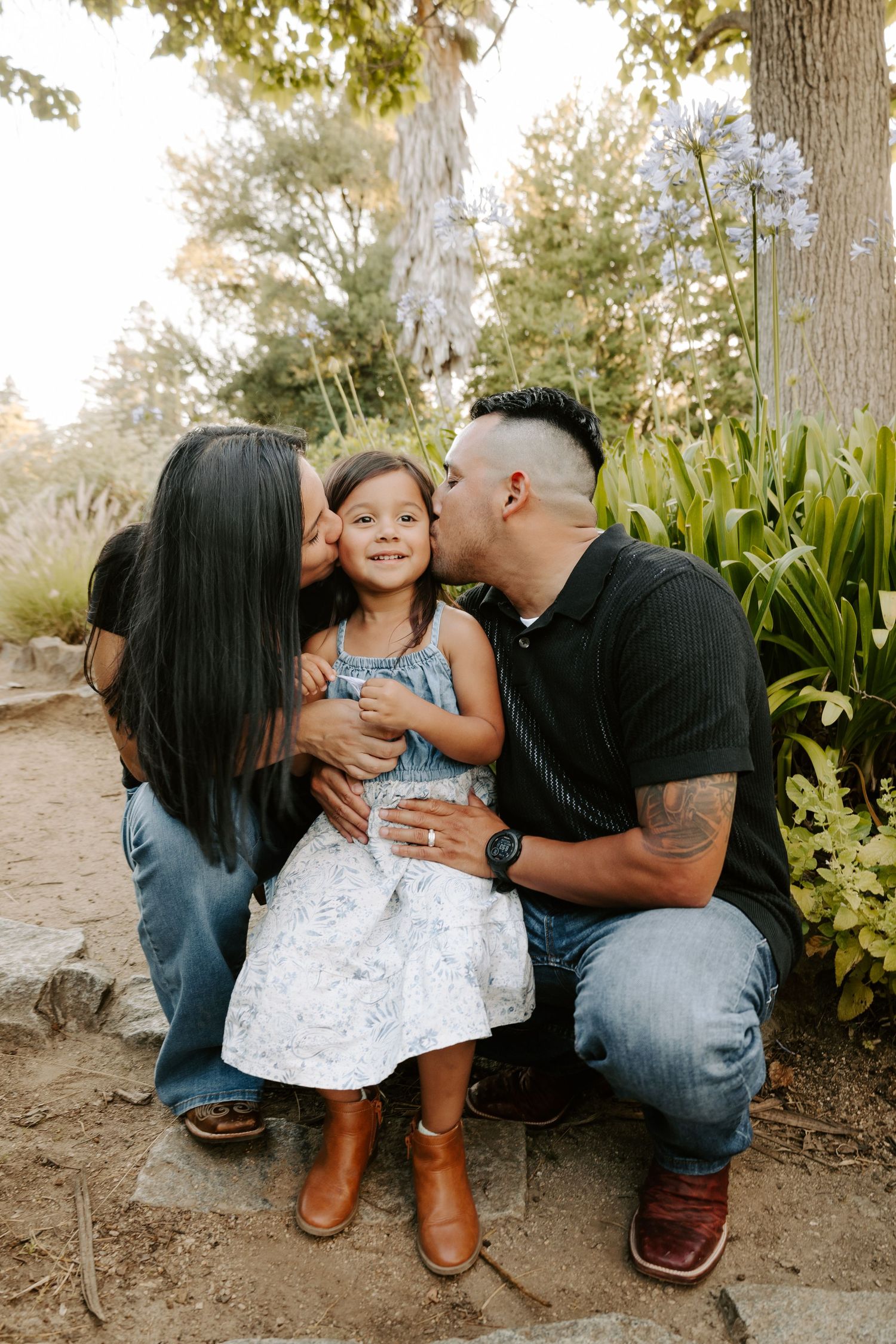 Parents kiss their smiling young daughter on the cheeks while sitting outdoors in a garden during a family portrait.