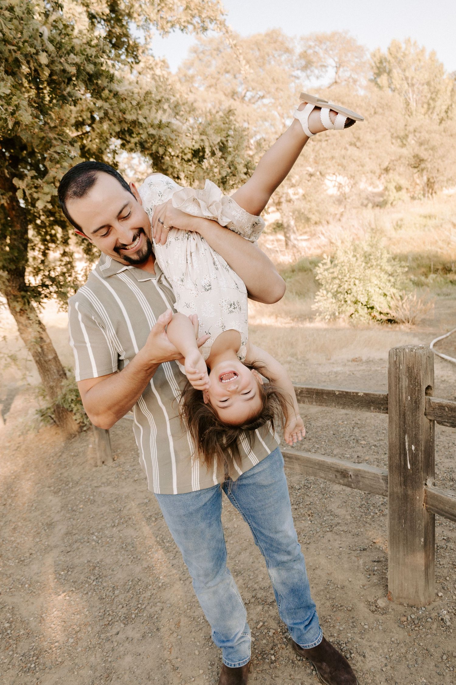 Laughing father playfully holds his young daughter upside down on a sunny dirt path during a family photoshoot.