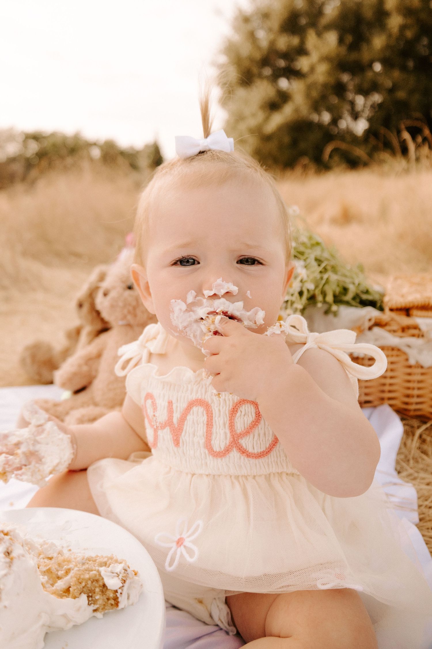 Adorable baby girl in white dress with one embroidery eating birthday cake at outdoor smash session.