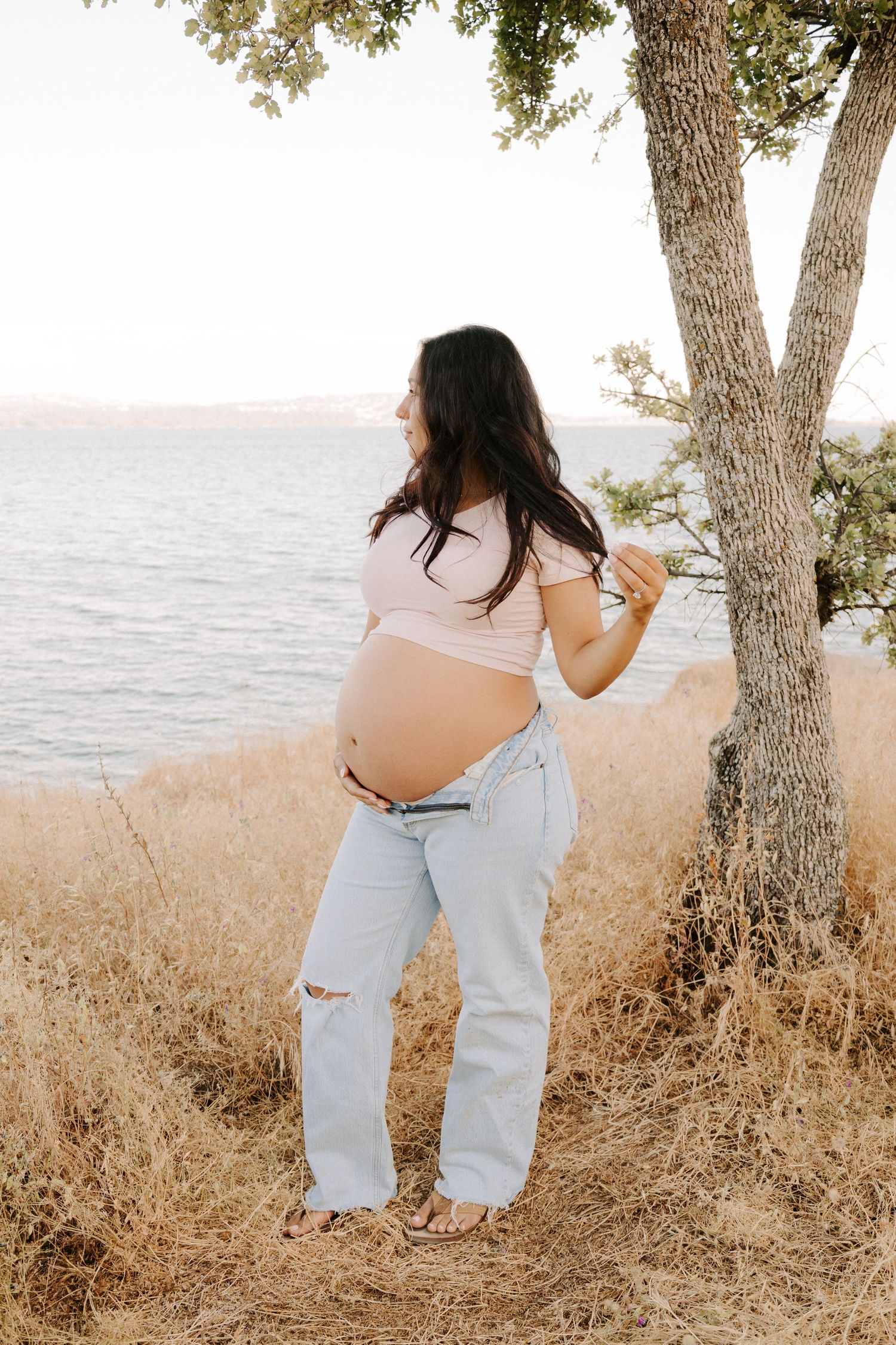 Pregnant woman in ripped jeans and pink top standing by a tree near a lake, cradling her baby bump outdoors.