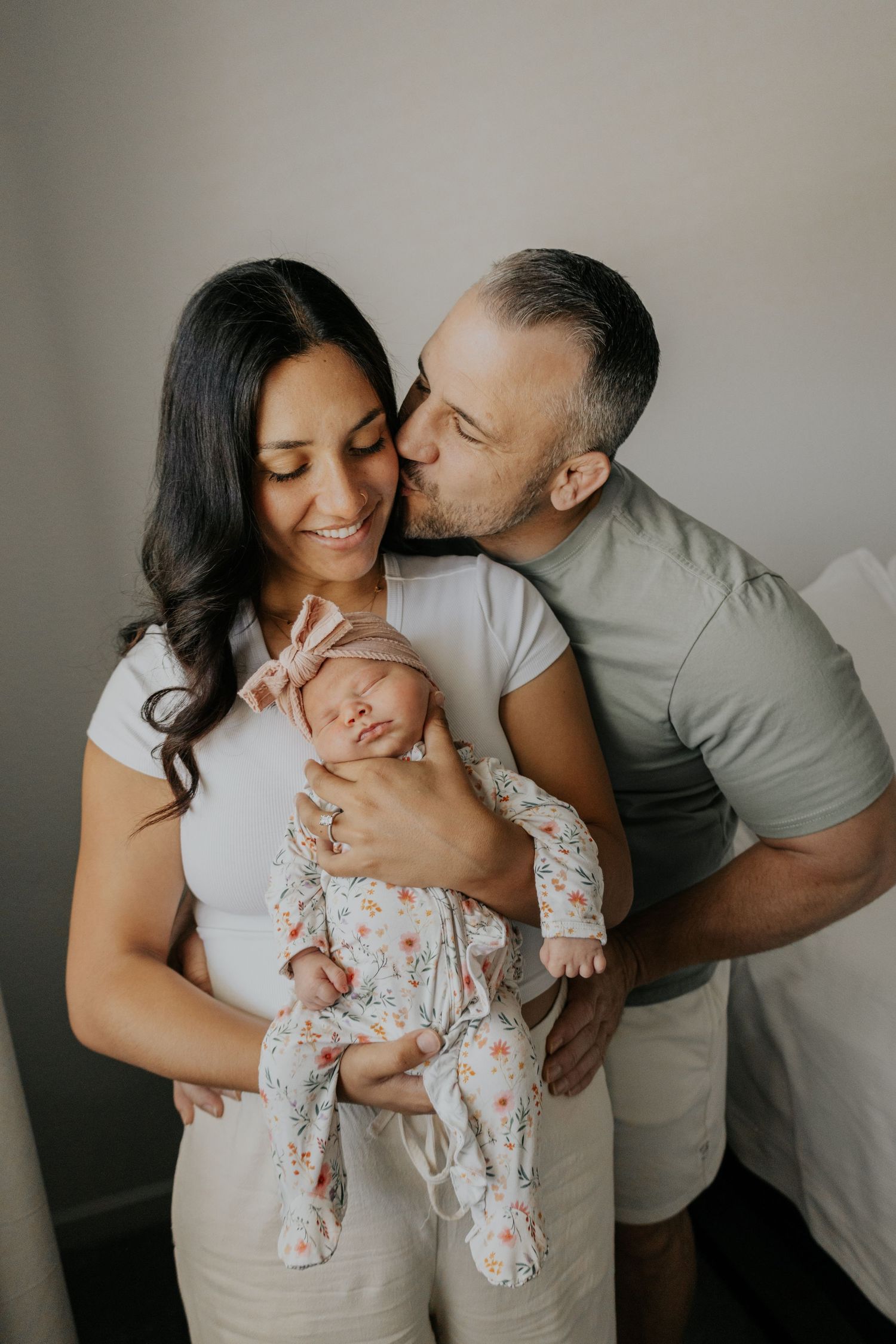 Happy couple with father kissing mothers head while she holds their sleeping newborn in floral outfit.
