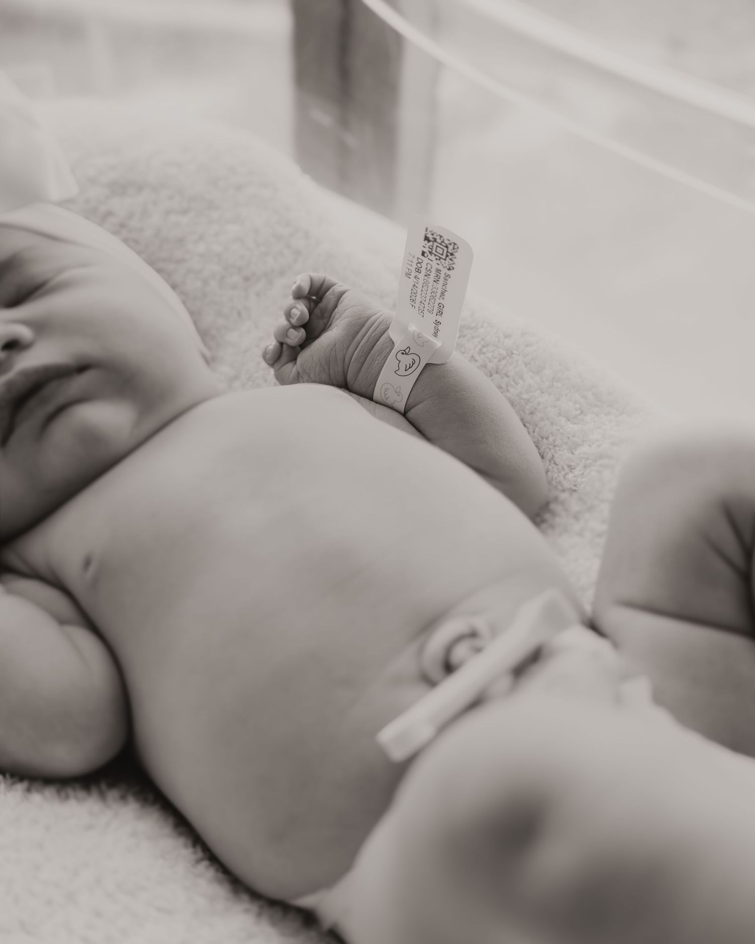 Peaceful newborn baby sleeping in hospital bassinet with identification bracelet, black and white photo.