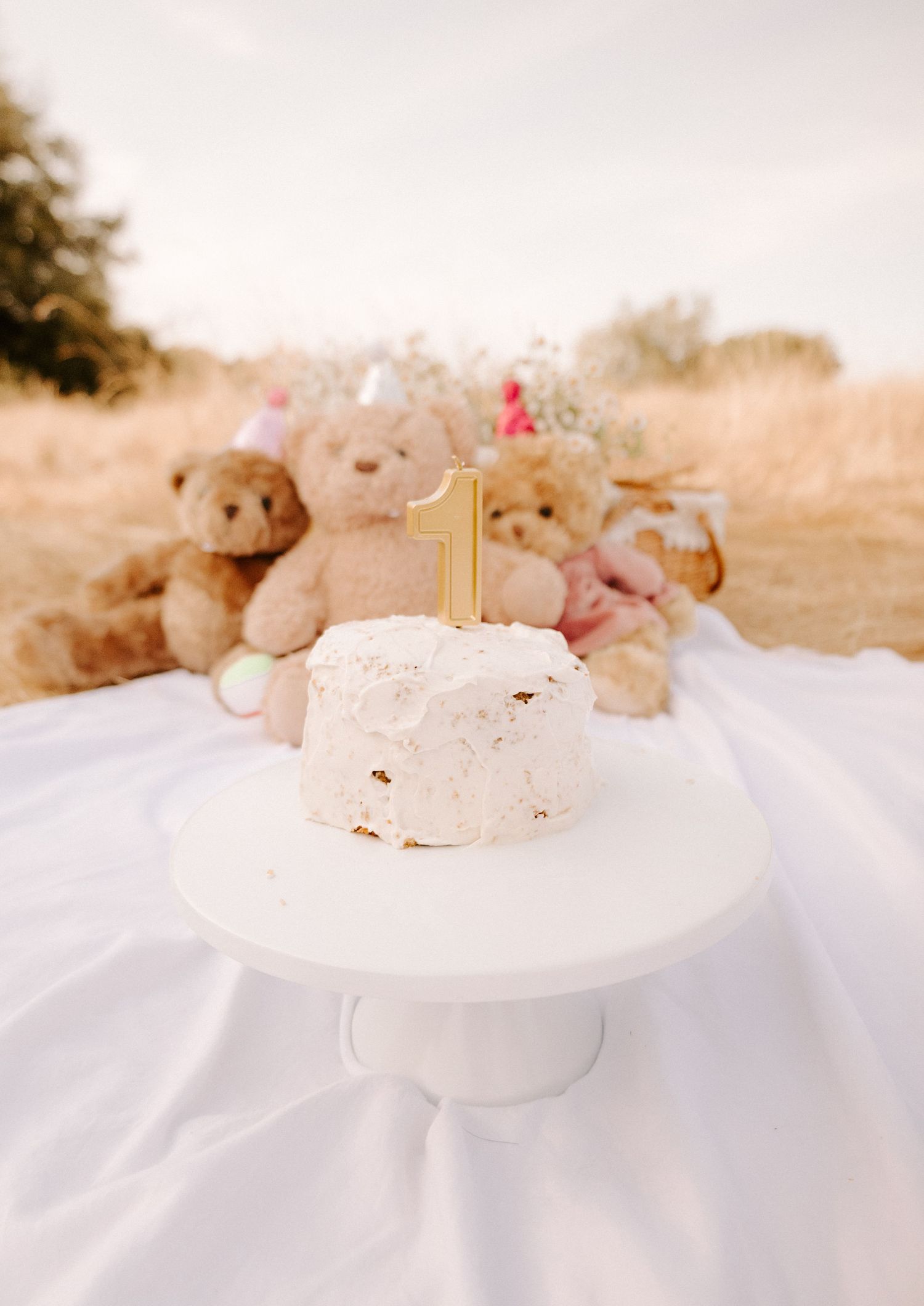 White first birthday smash cake with gold number one topper on cake stand with teddy bears in field.