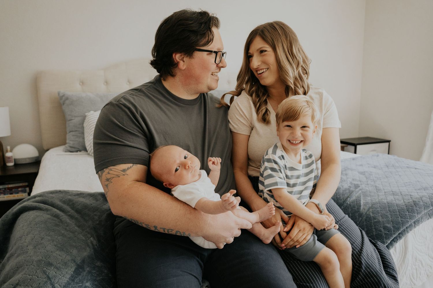 Happy family of four sitting on bed with newborn and toddler smiling together.