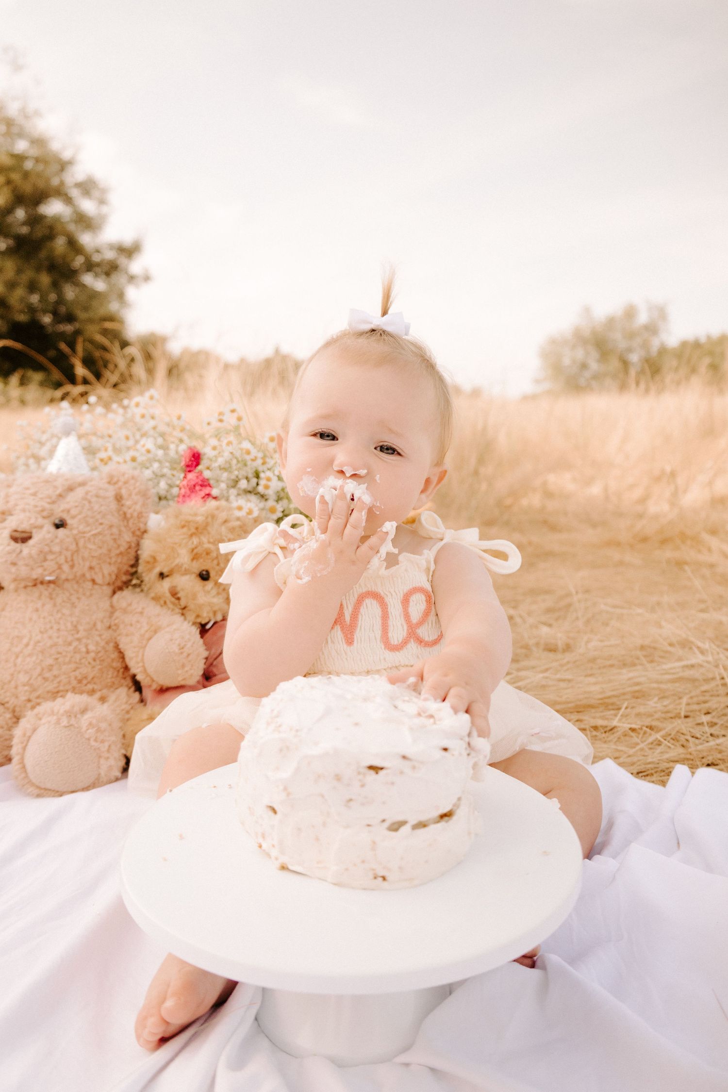 Baby girl celebrating first birthday smashing white cake outdoors in golden field.