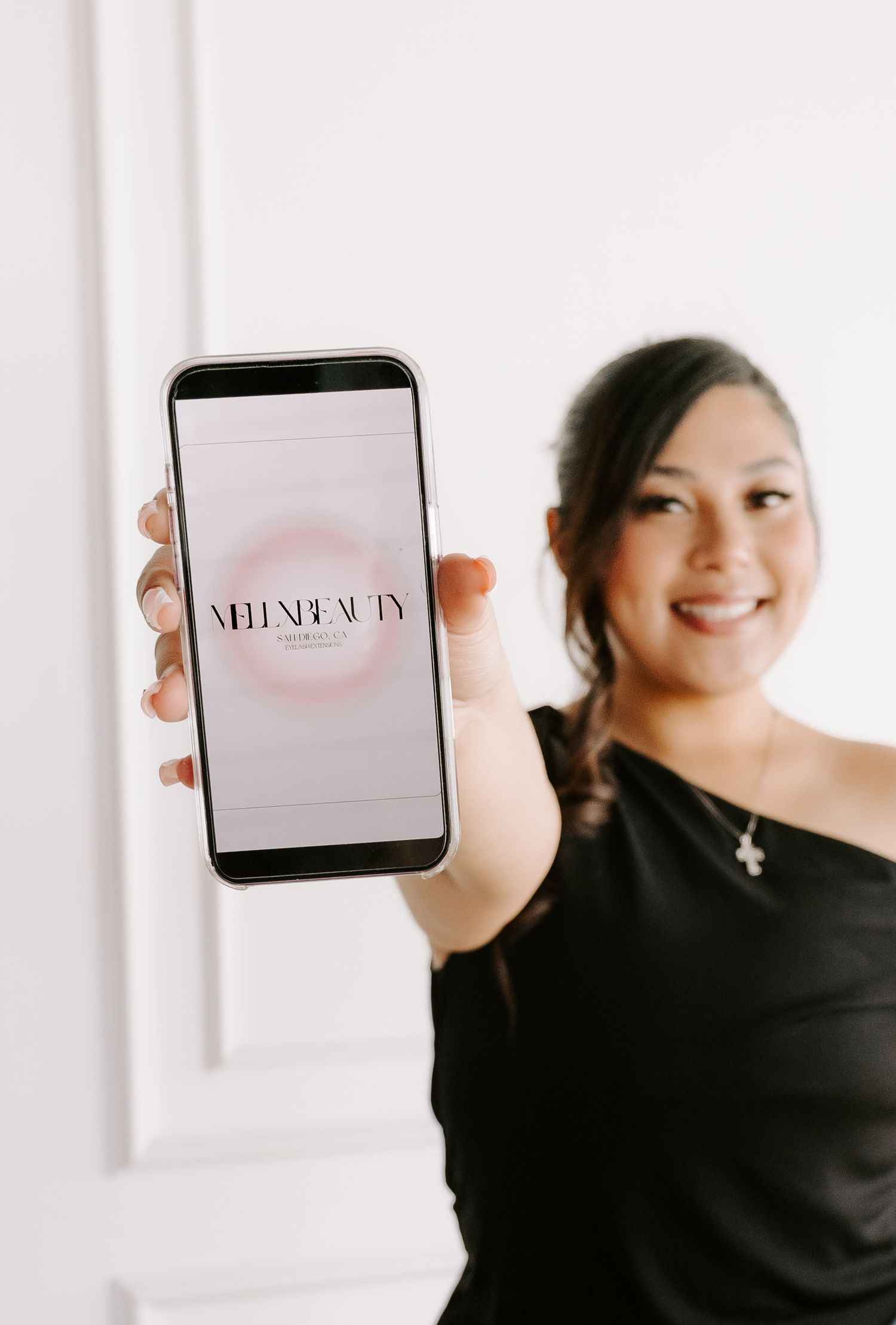 Woman in black top  smiling and holding phone displaying MelxBeauty app logo against white background.