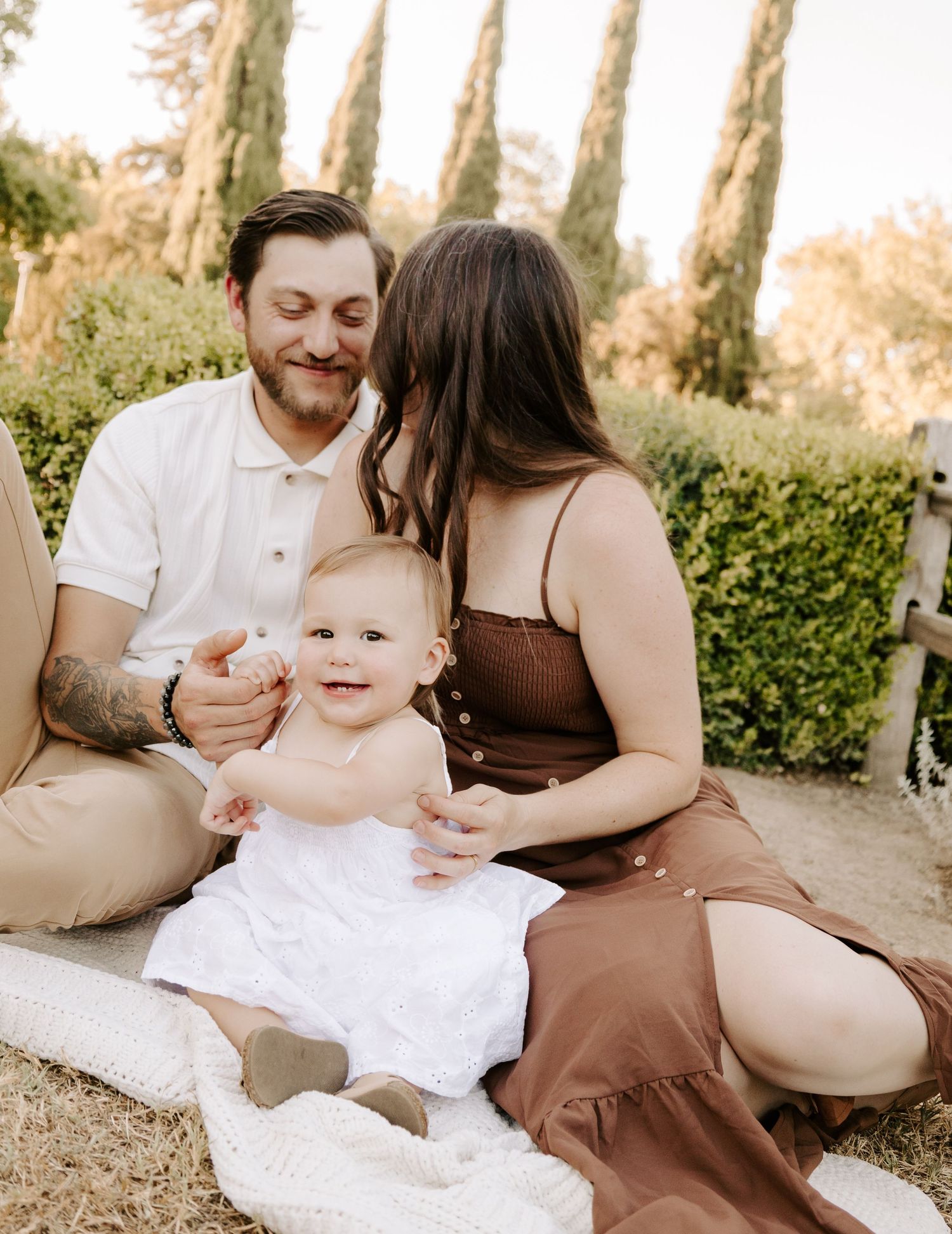 Young parents sitting on a blanket outdoors, lovingly attending to their smiling baby girl.