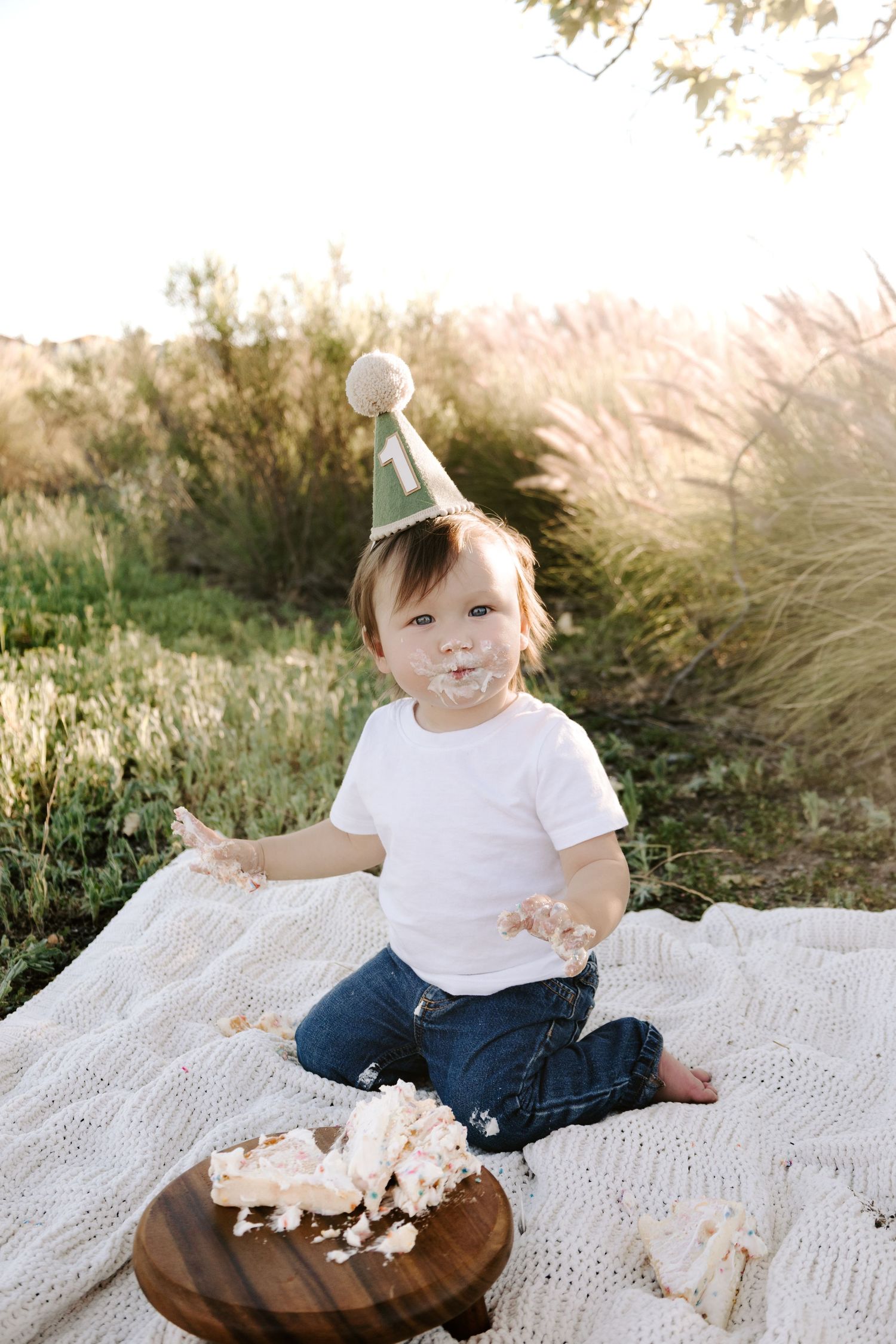 Toddler in birthday hat smashing first cake outdoors on blanket in sunny field.