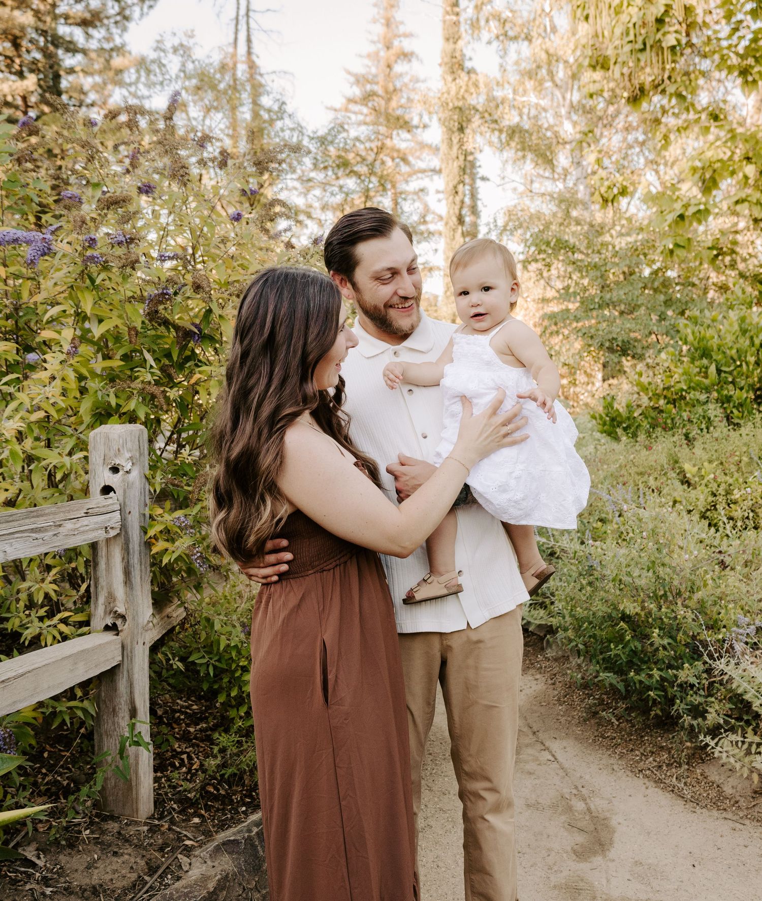 A smiling couple stands on a forest path, with the man holding a happy baby in a white outfit.