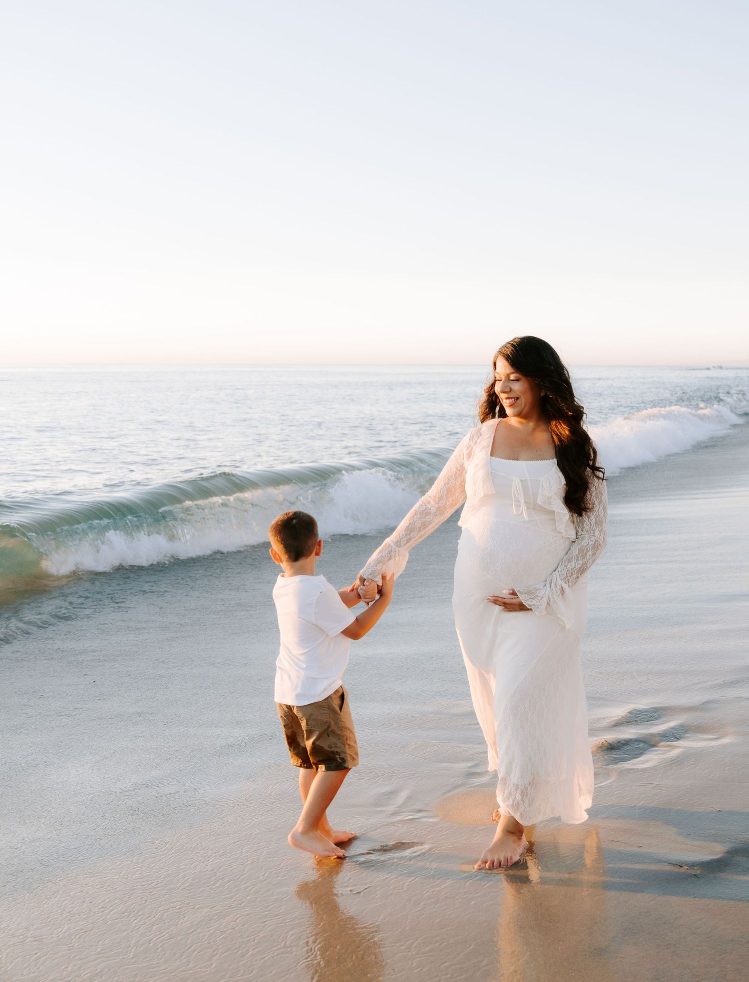 Smiling pregnant woman in white dress holds hands with young son on sunny beach.