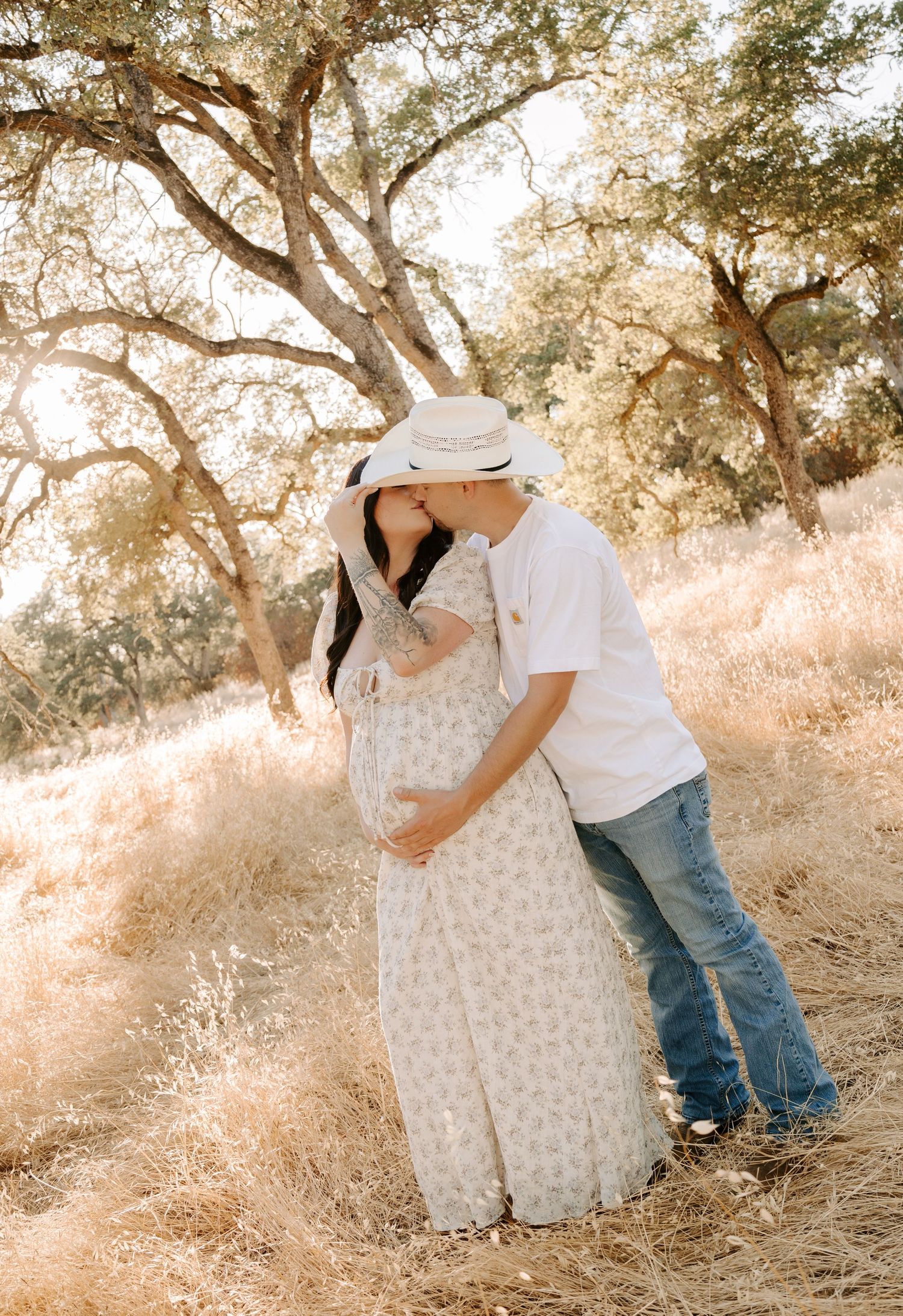Couple shares a kiss in a sunlit golden field, man in white hat and jeans, woman in floral maternity dress.