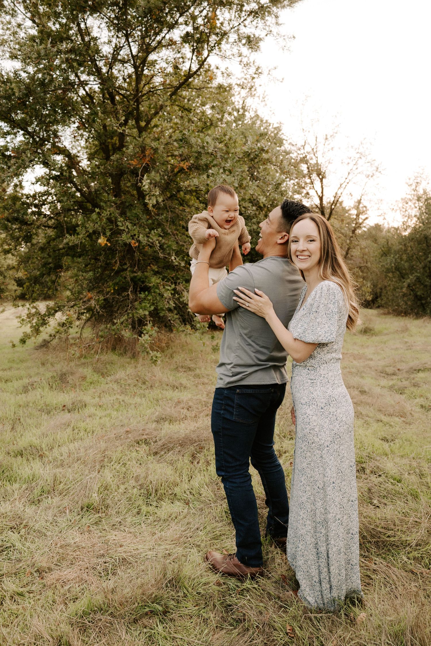 Smiling family of three poses in a green meadow as father lifts laughing baby while mother stands beside them.