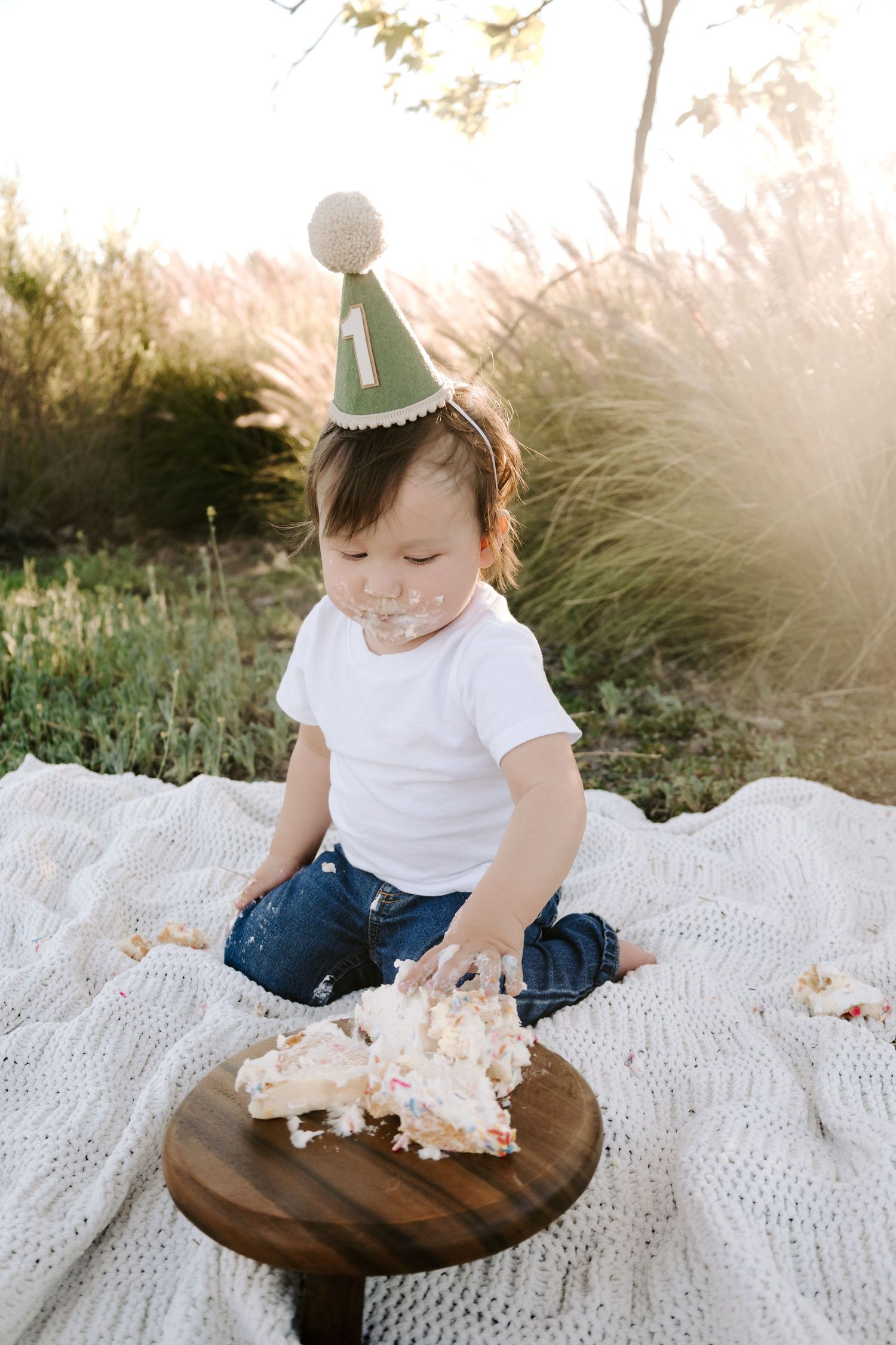 A baby wearing a green number one birthday hat smashes their first birthday cake outdoors at sunset.
