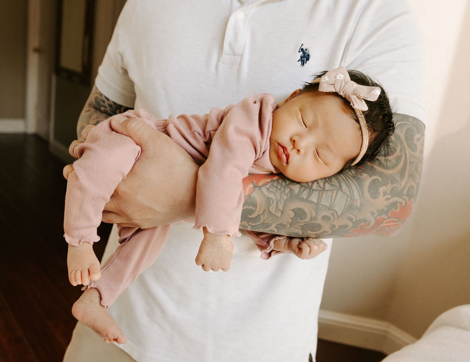 Sleeping newborn baby girl in pink outfit resting on fathers tattooed arm with bow headband.