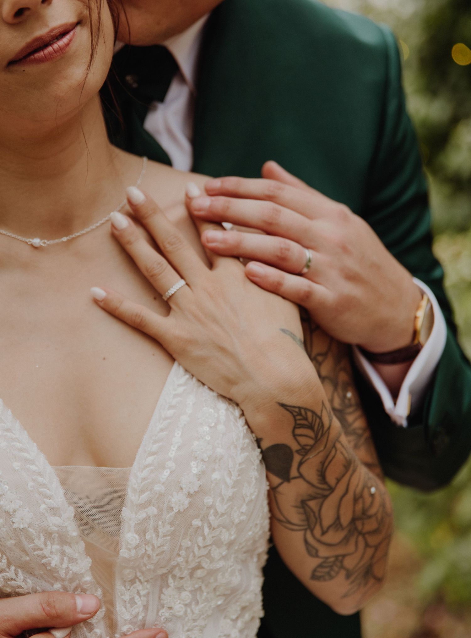Gros plan sur des alliances et des mains ornées d'un tatouage floral sur fond naturel flou.