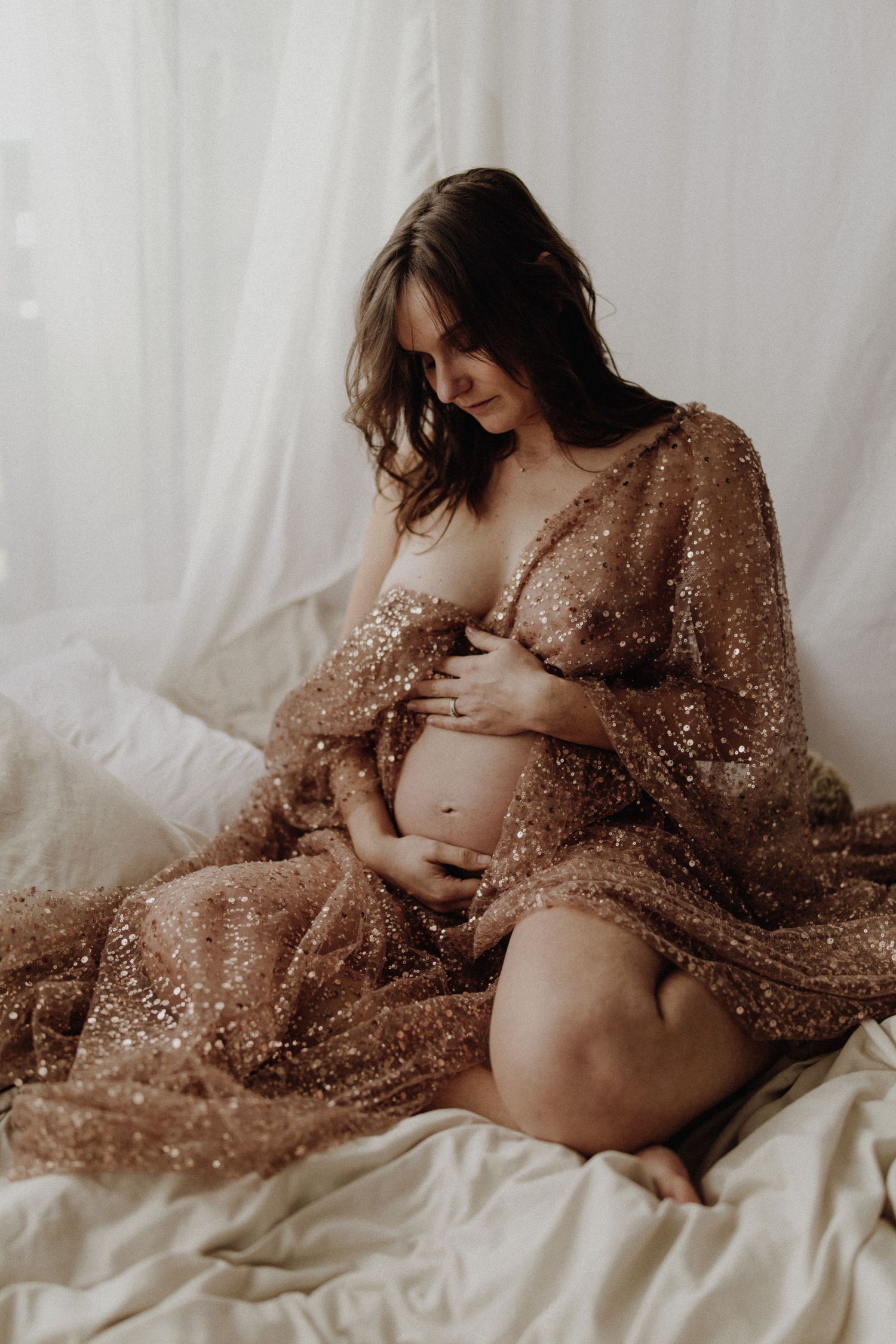 Séance photo artistique de maternité mettant en scène une future maman vêtue d'une robe bronze scintillante, allongée sur un fond blanc.