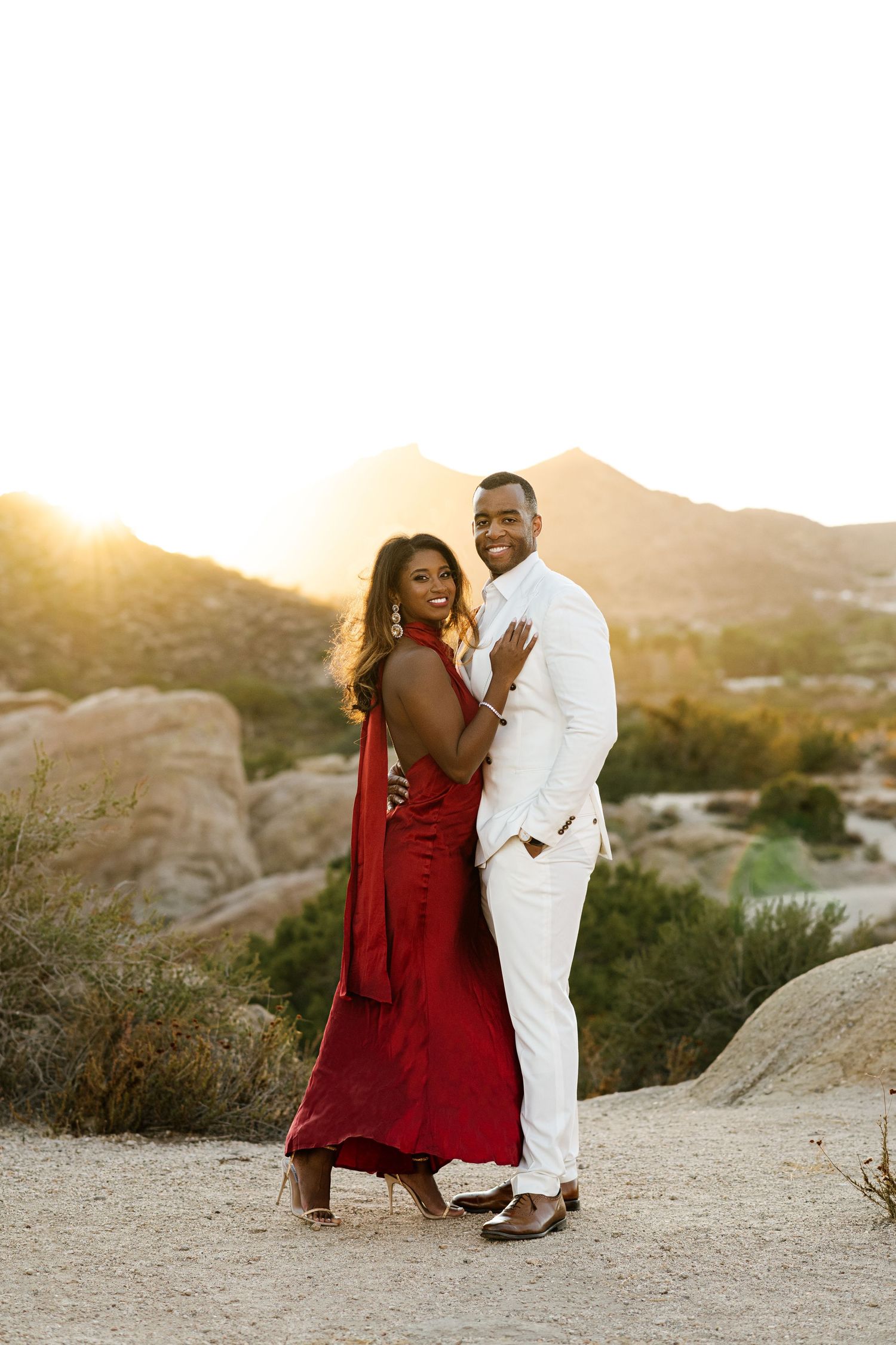 Couple in red dress and white suit pose for romantic desert photoshoot at sunset.