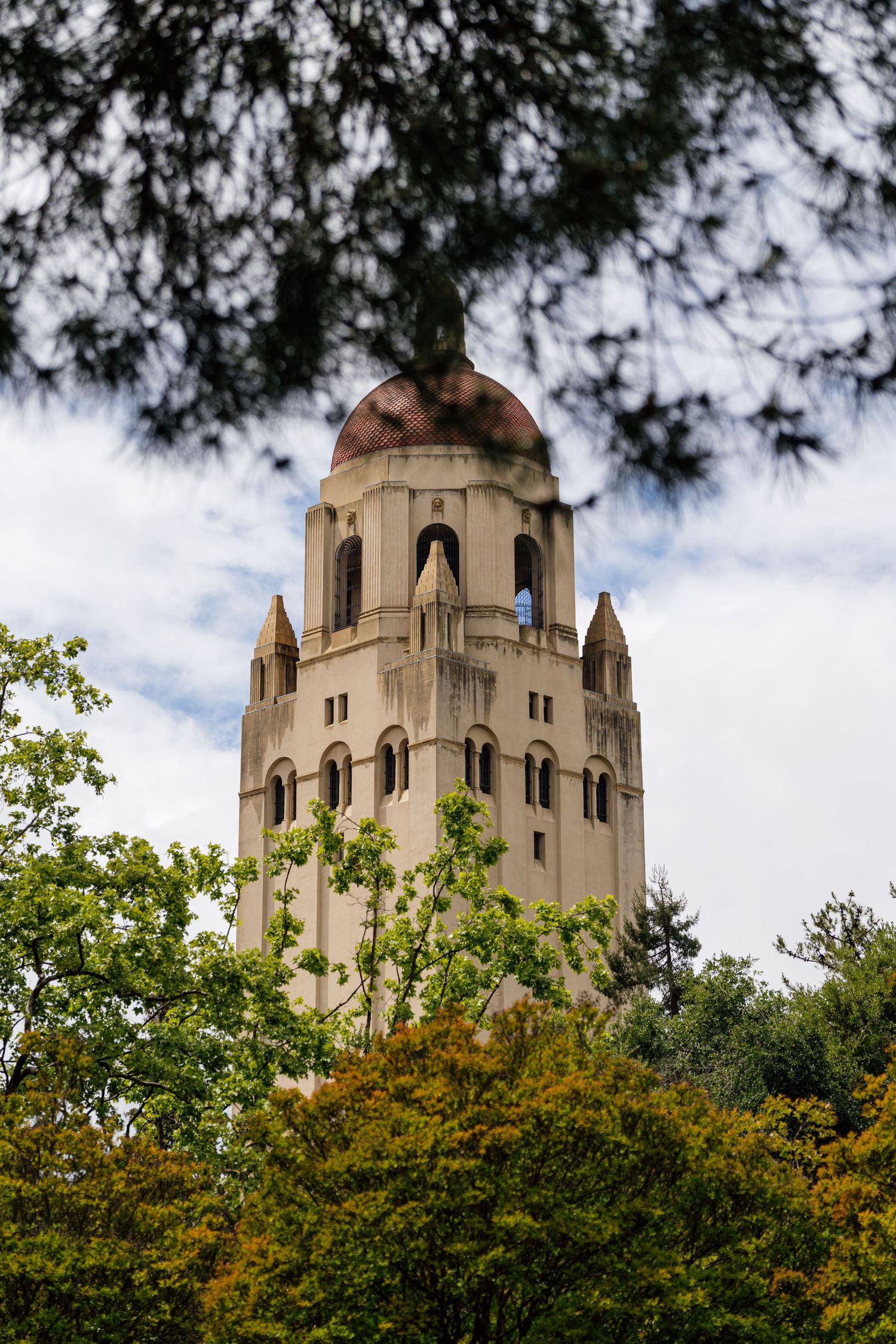Hoover Tower at Stanford University rises above lush green trees under a cloudy sky.