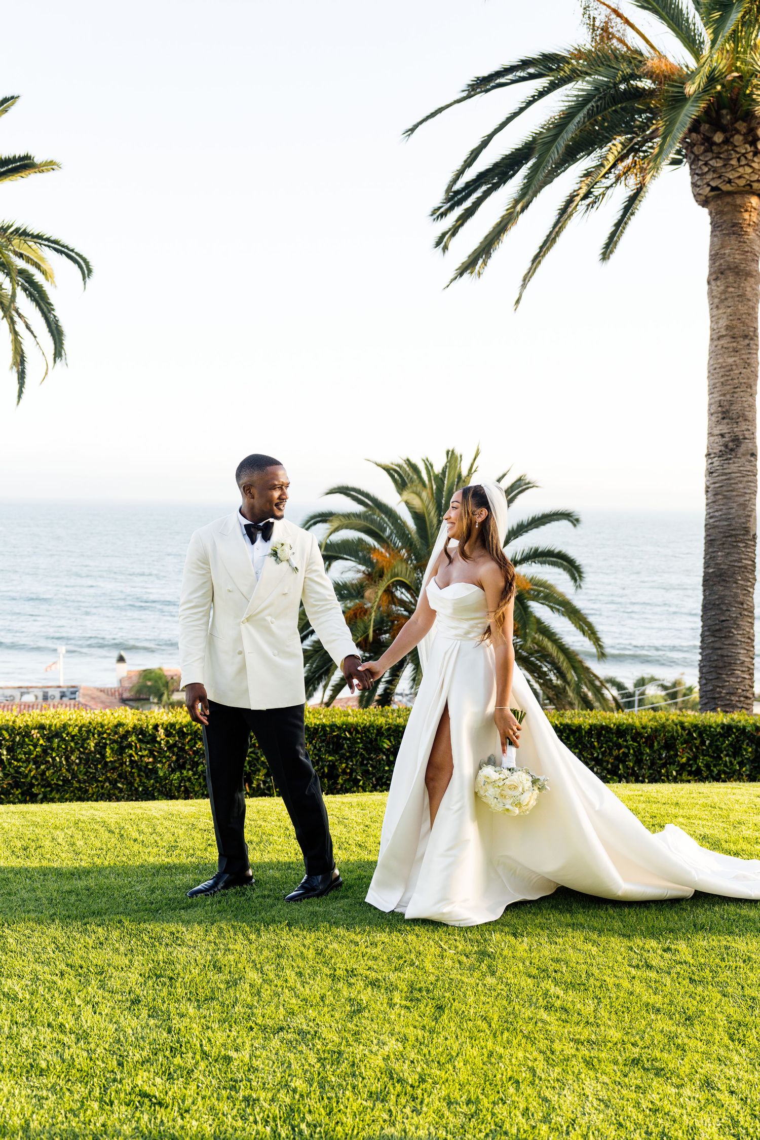 A bride and groom hold hands on a green lawn with palm trees and ocean views at a sunny outdoor wedding.