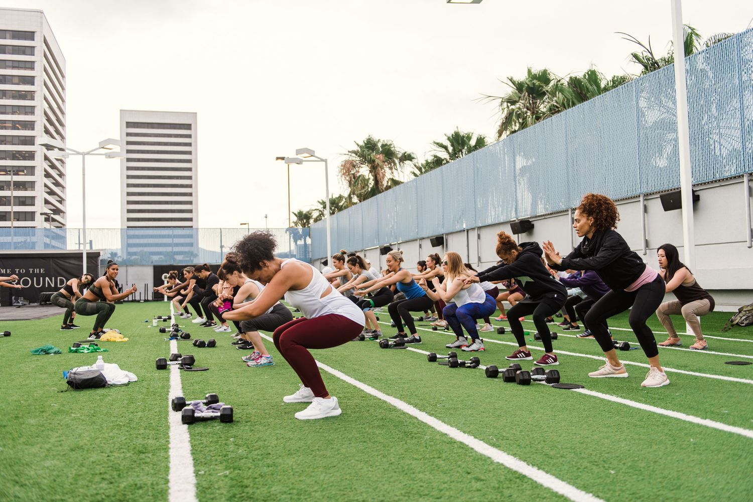 Group fitness class doing squats outdoors on artificial turf with urban backdrop.