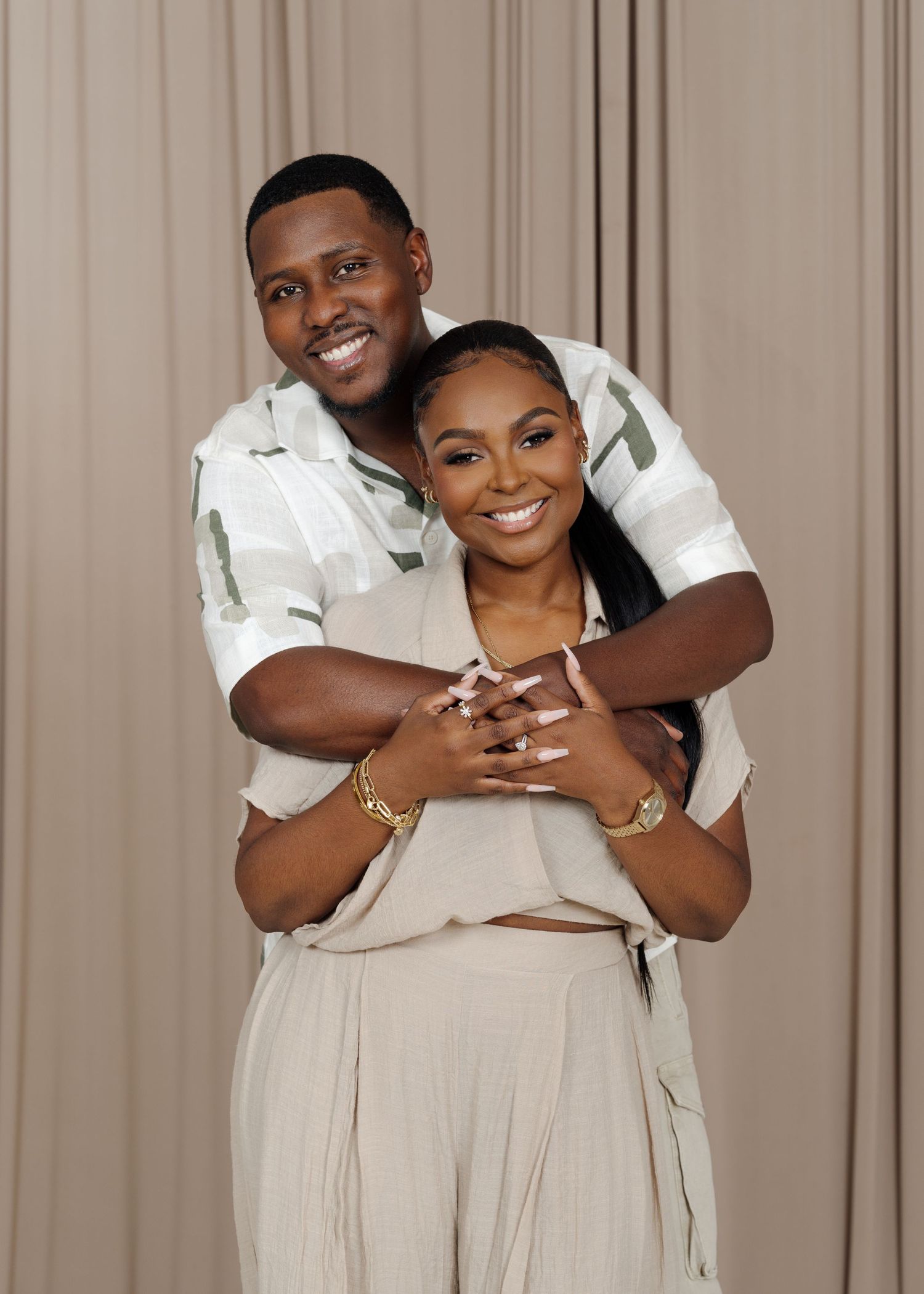 Happy couple embraces and laughs together in matching cream-colored outfits against a neutral backdrop.