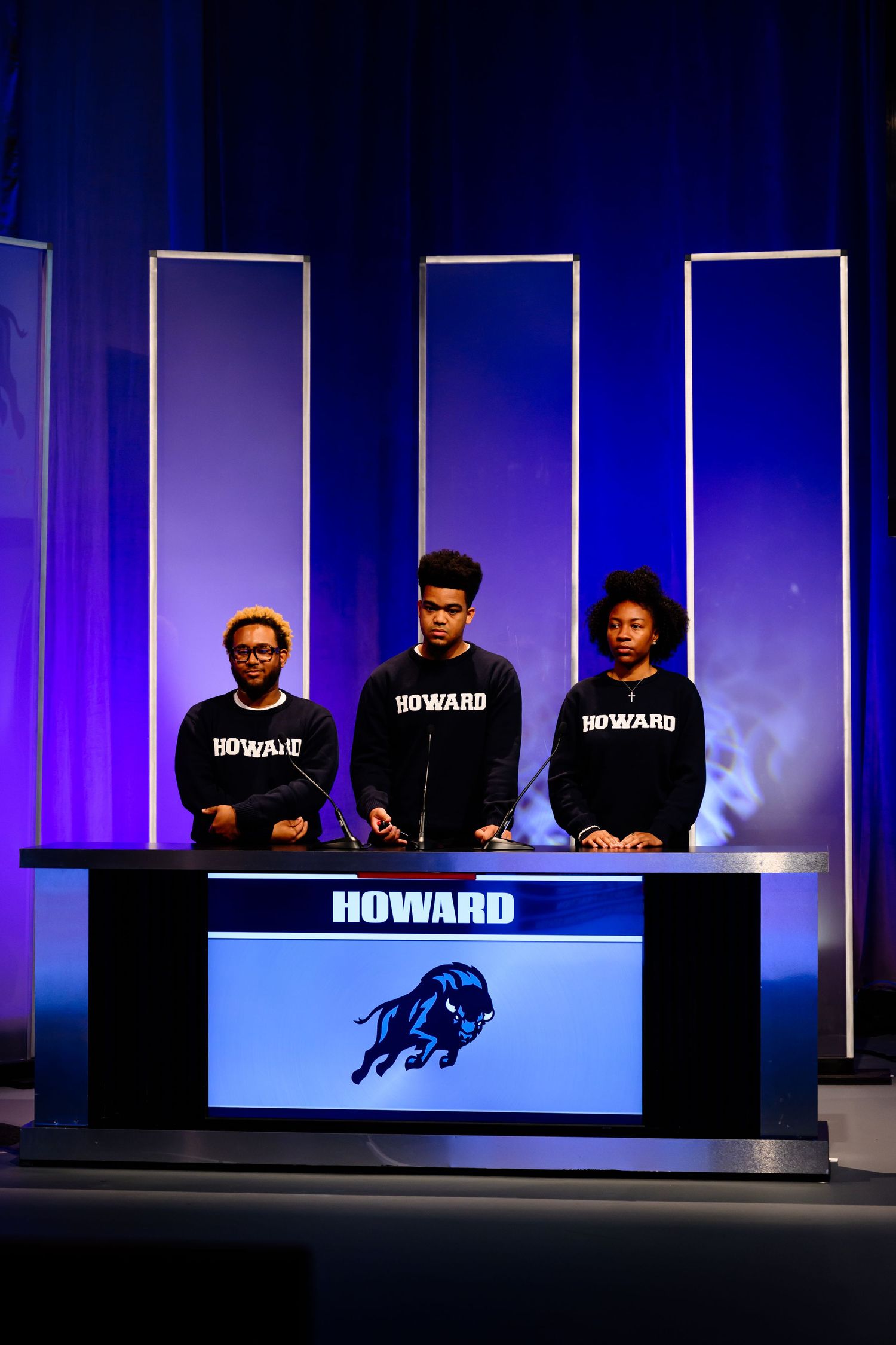 Contestants in matching black shirts seated at competition podiums with blue stage lighting.