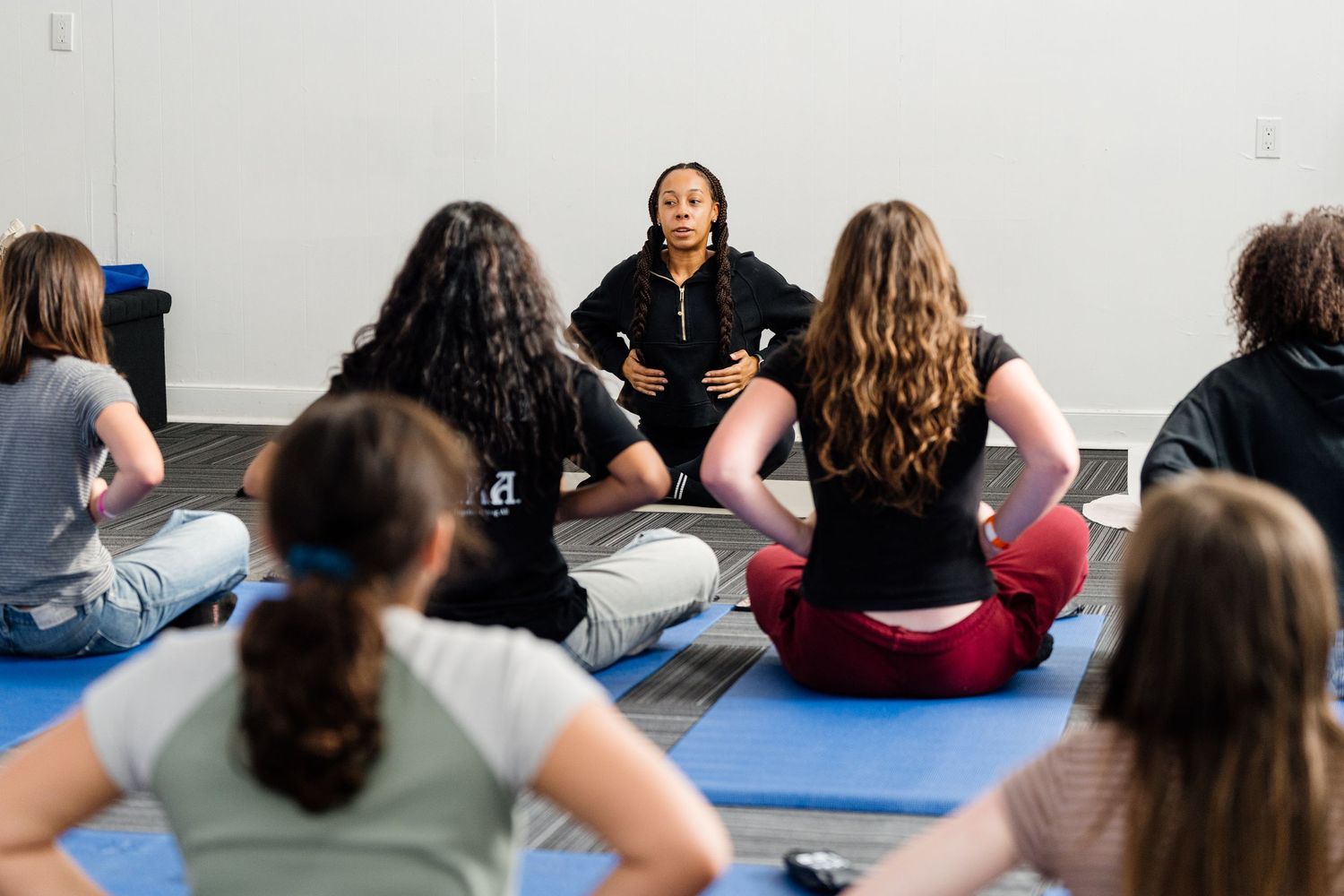 Yoga instructor leads a seated meditation class with students on blue mats in a bright studio.