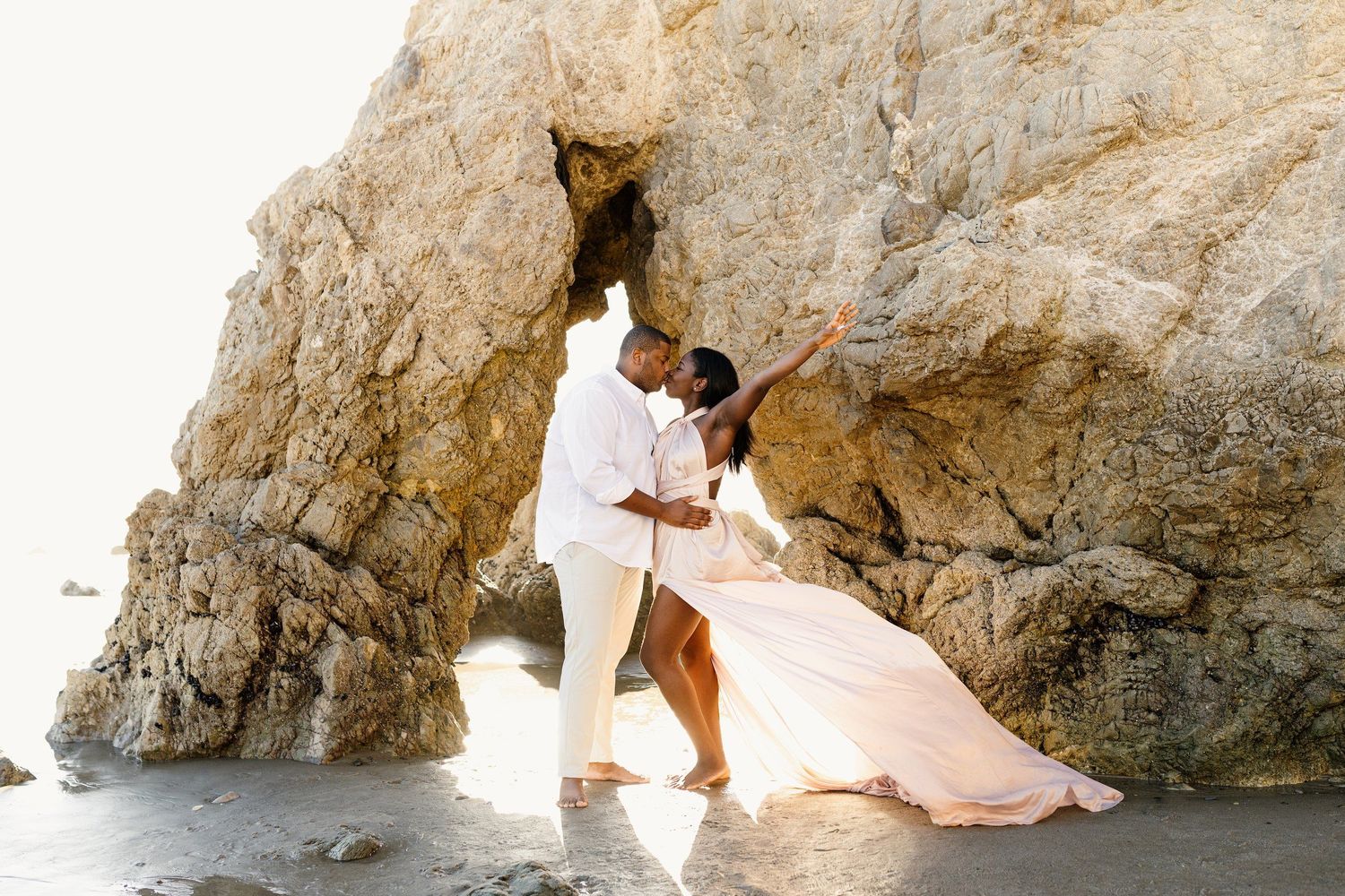 Couple in flowing white outfits poses romantically by coastal rock formation.