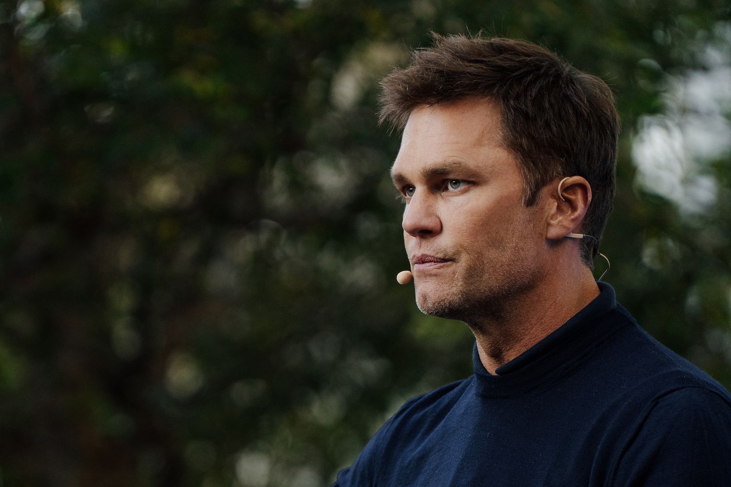 A person in a navy blue turtleneck sweater gazes thoughtfully into the distance against a blurred outdoor background.