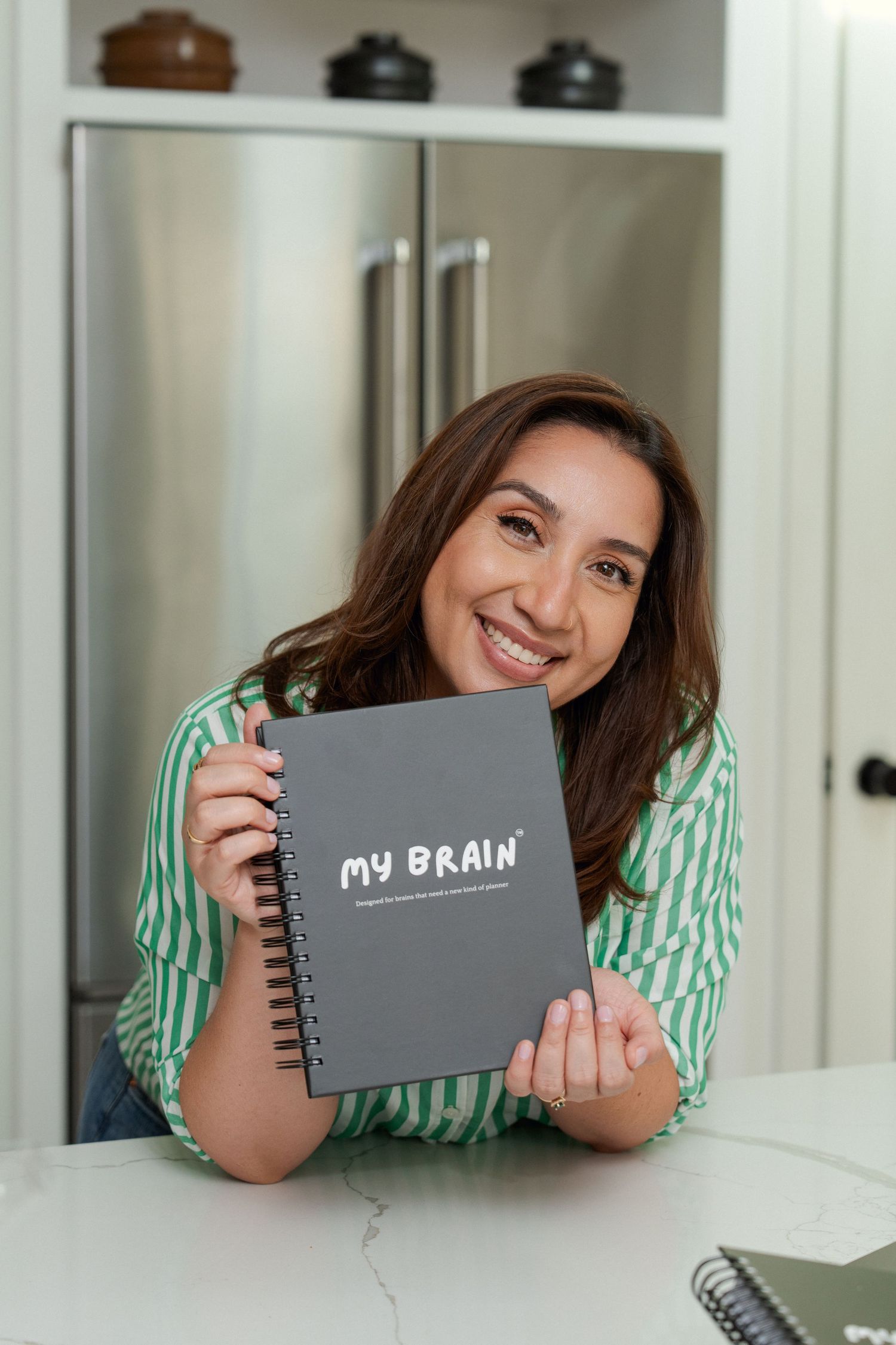 Smiling woman holding a gray spiral notebook labeled My Brain in a modern kitchen.
