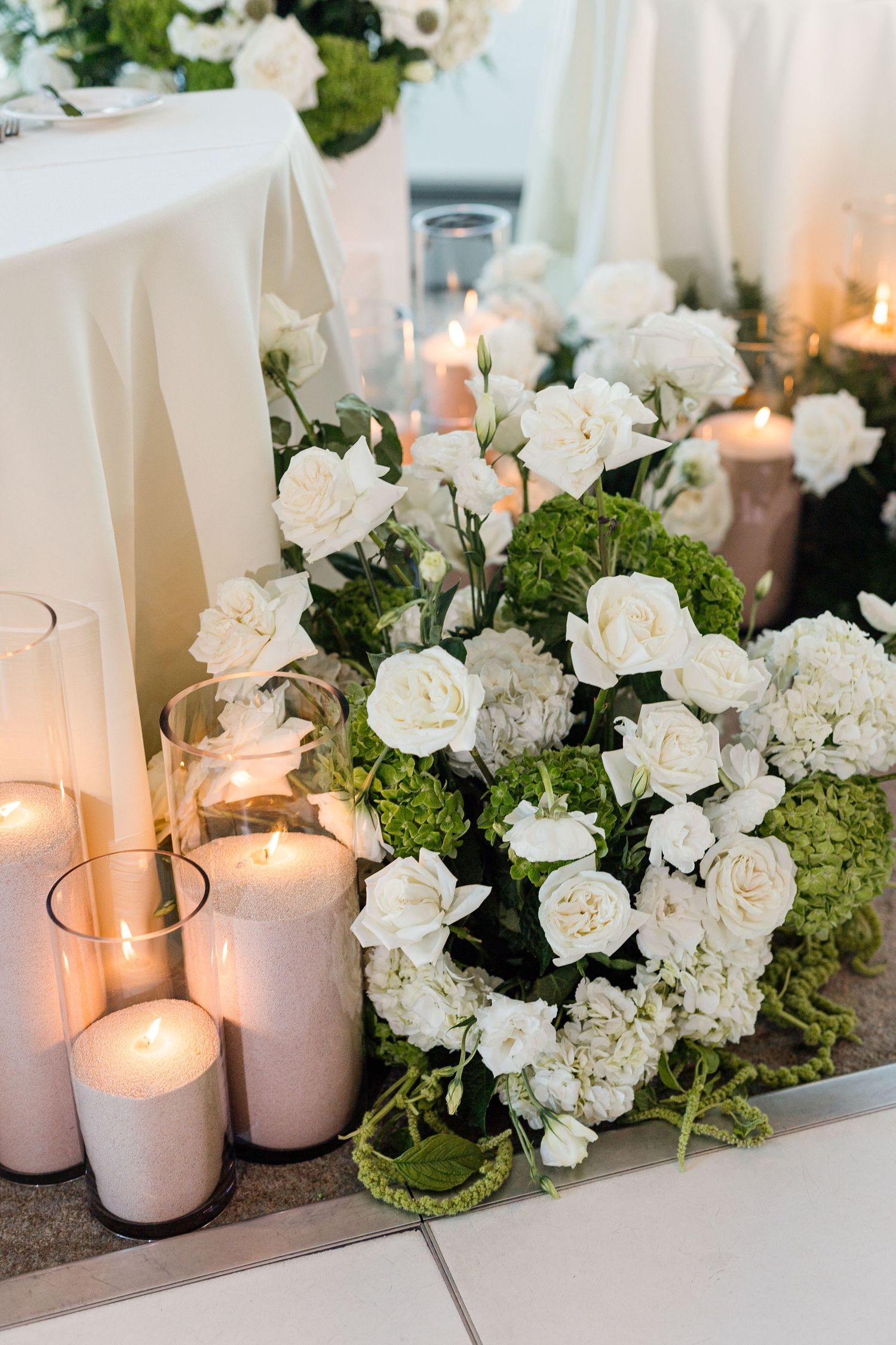 Elegant wedding floral arrangement with white roses, green hydrangeas, and lit pillar candles on a white tablecloth.