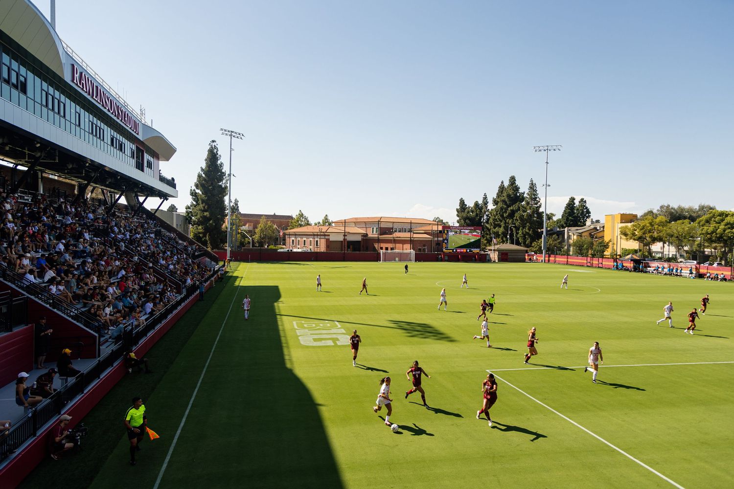 Wide view of a soccer stadium during daytime with players on the field and spectators in stands.