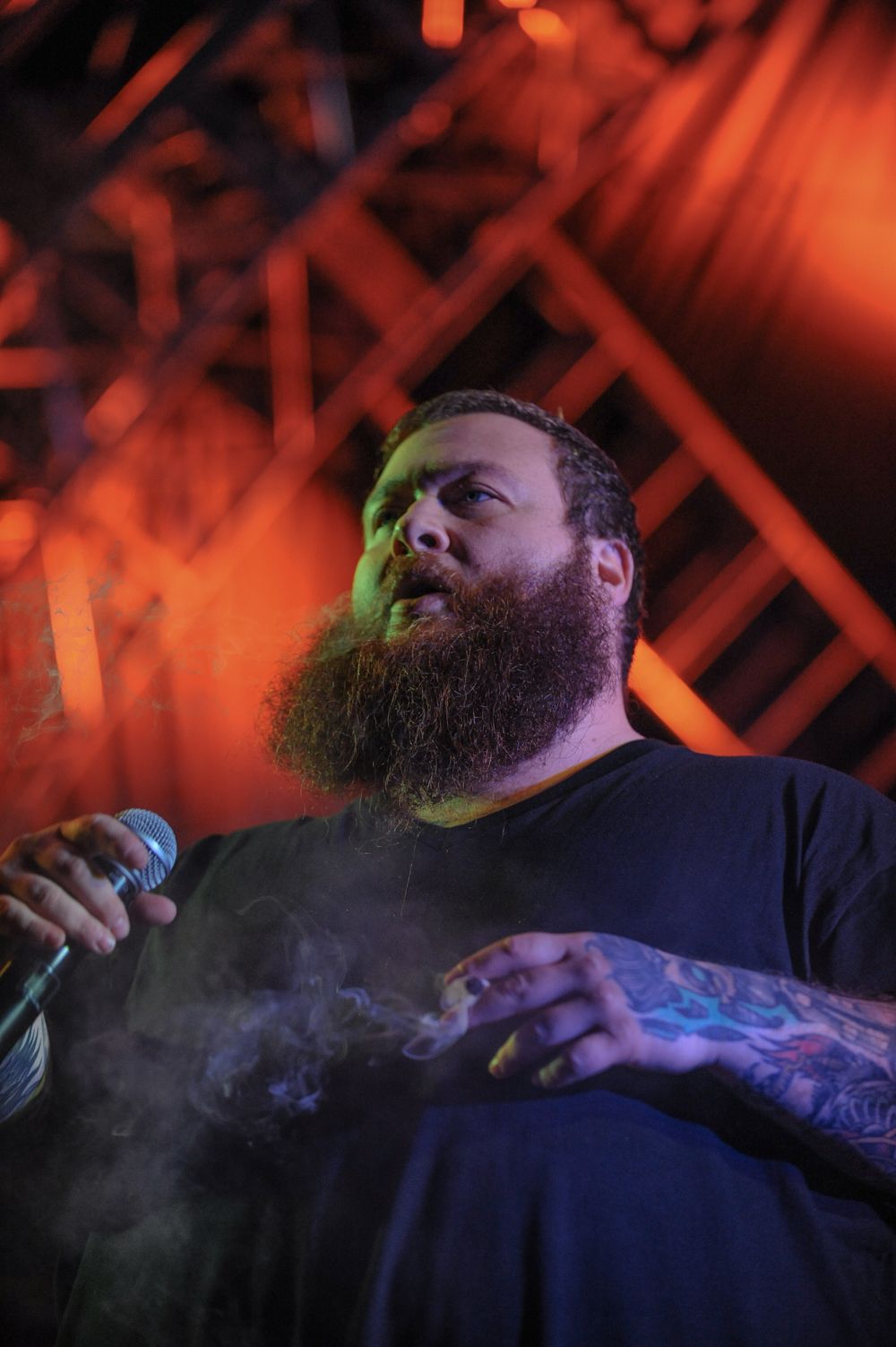 Performer with a full beard singing in dramatic orange lighting on stage.