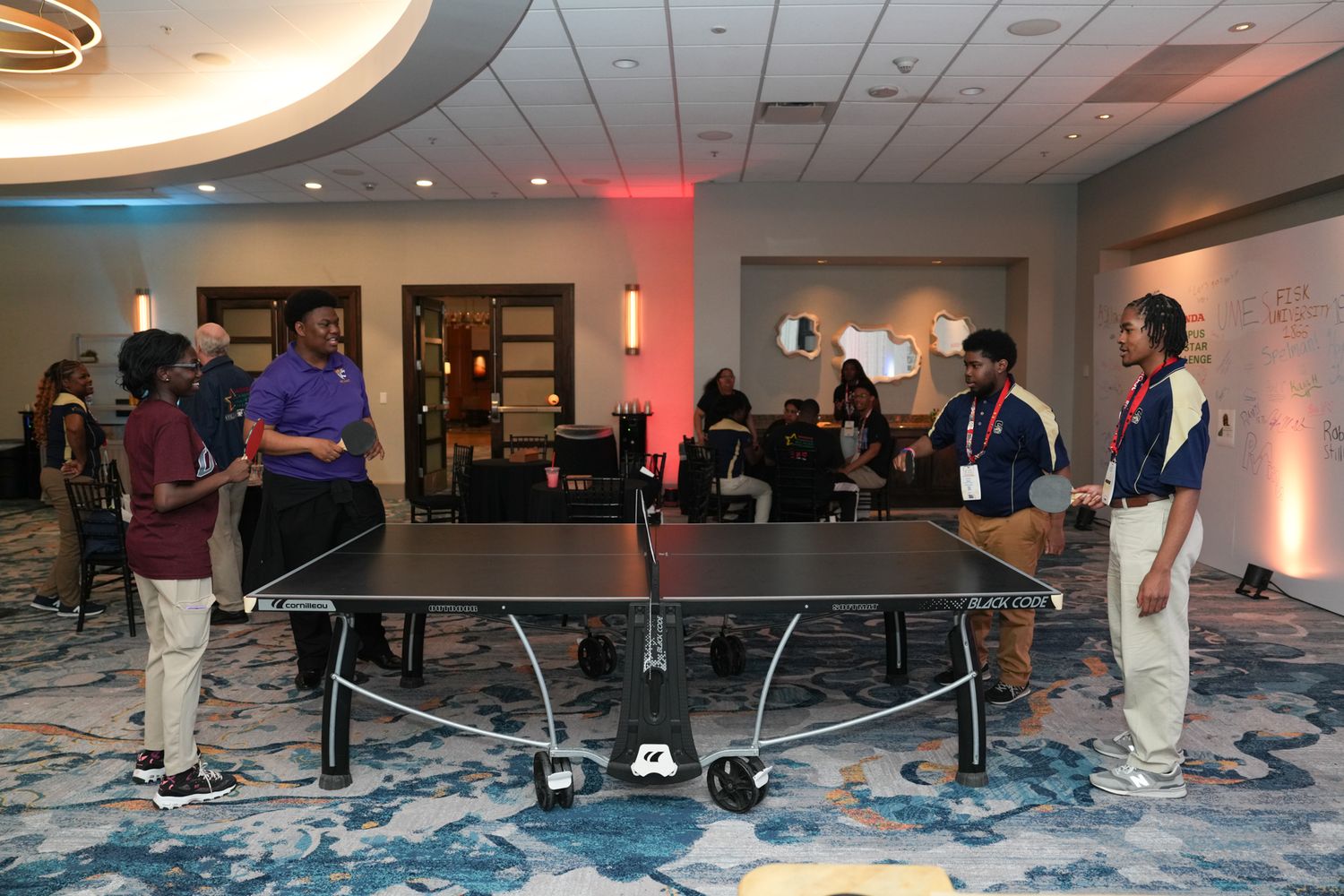 People playing ping pong at an indoor event in a hotel ballroom with colorful lighting and guests mingling.